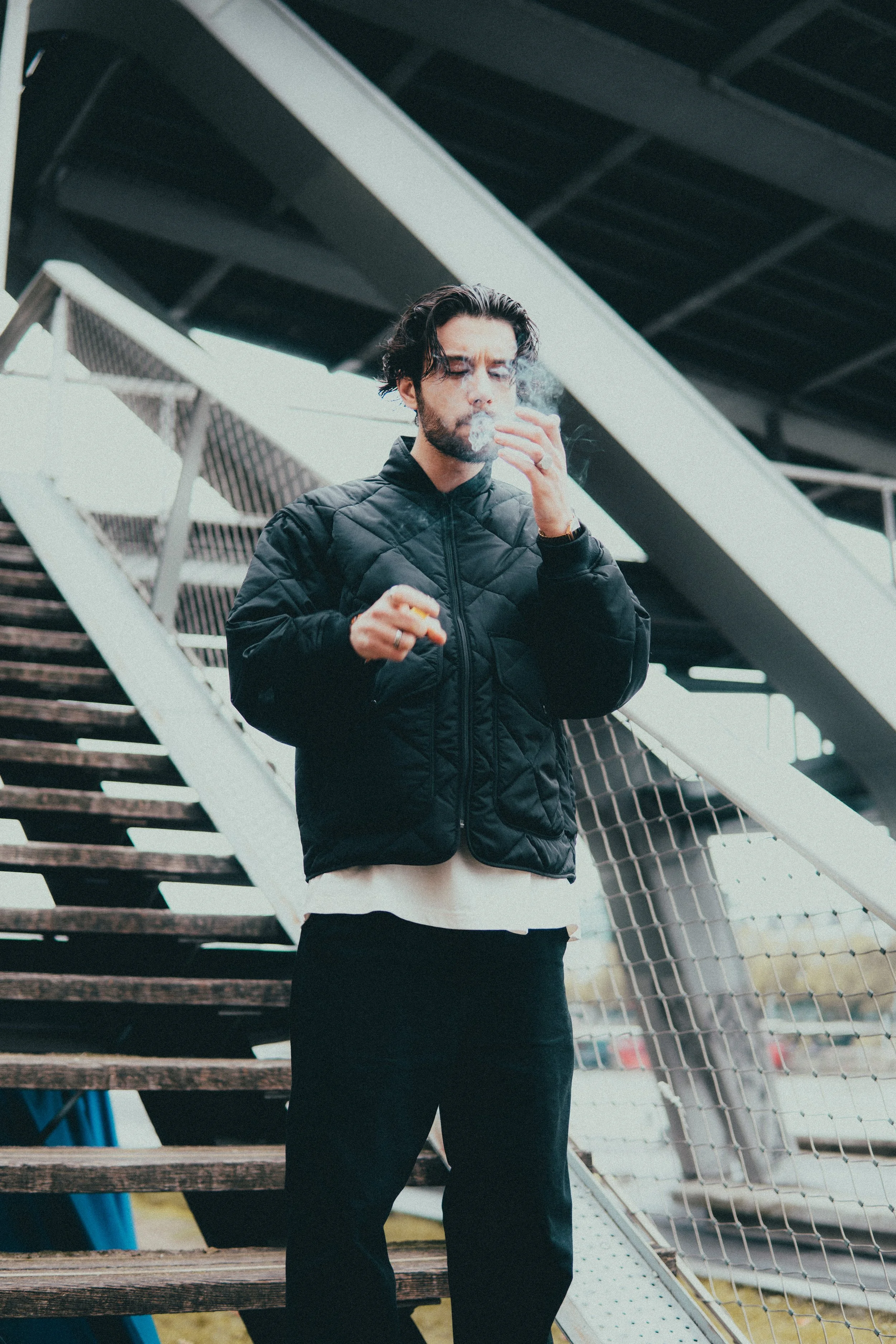 Un homme noir portant une veste noire et un pantalon noir, qui fume une cigarette, se tient près d'un escalier en bois et en métal en extérieur.