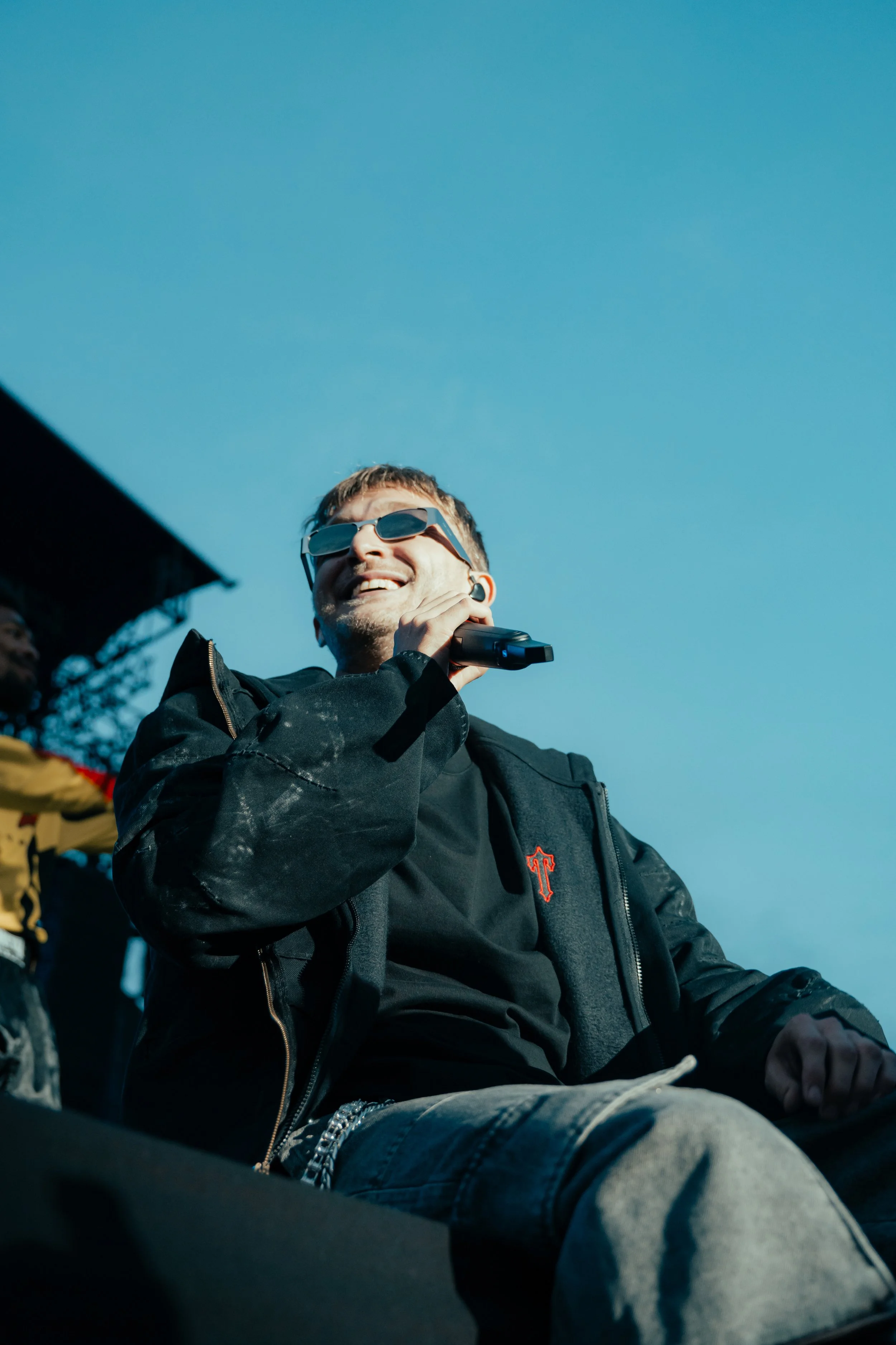 Un homme souriant portant des lunettes de soleil, une veste noire avec une broderie rouge, et une chaîne, assis en plein air sous un ciel bleu, tenant un microphone ou un objet à la main.