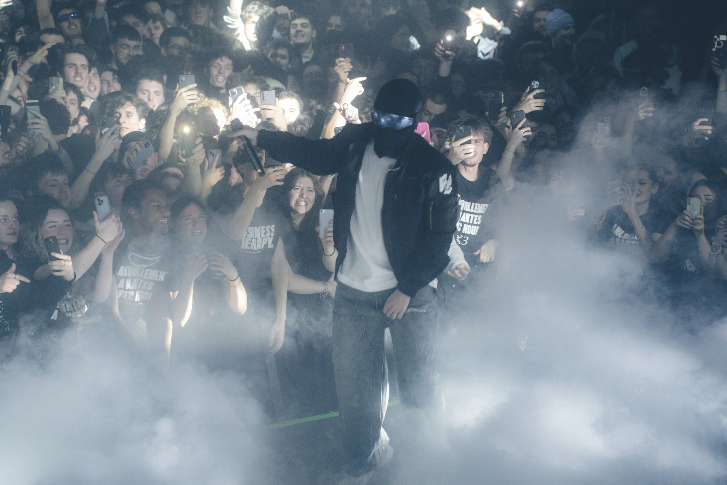 Un homme avec un masque noir, veste en cuir et pantalon décontracté se tient au centre d'une foule de jeunes, prenant des photos ou vidéos avec leurs téléphones portables, dans une atmosphère dynamique avec de la fumée et une forte lumière en arrière