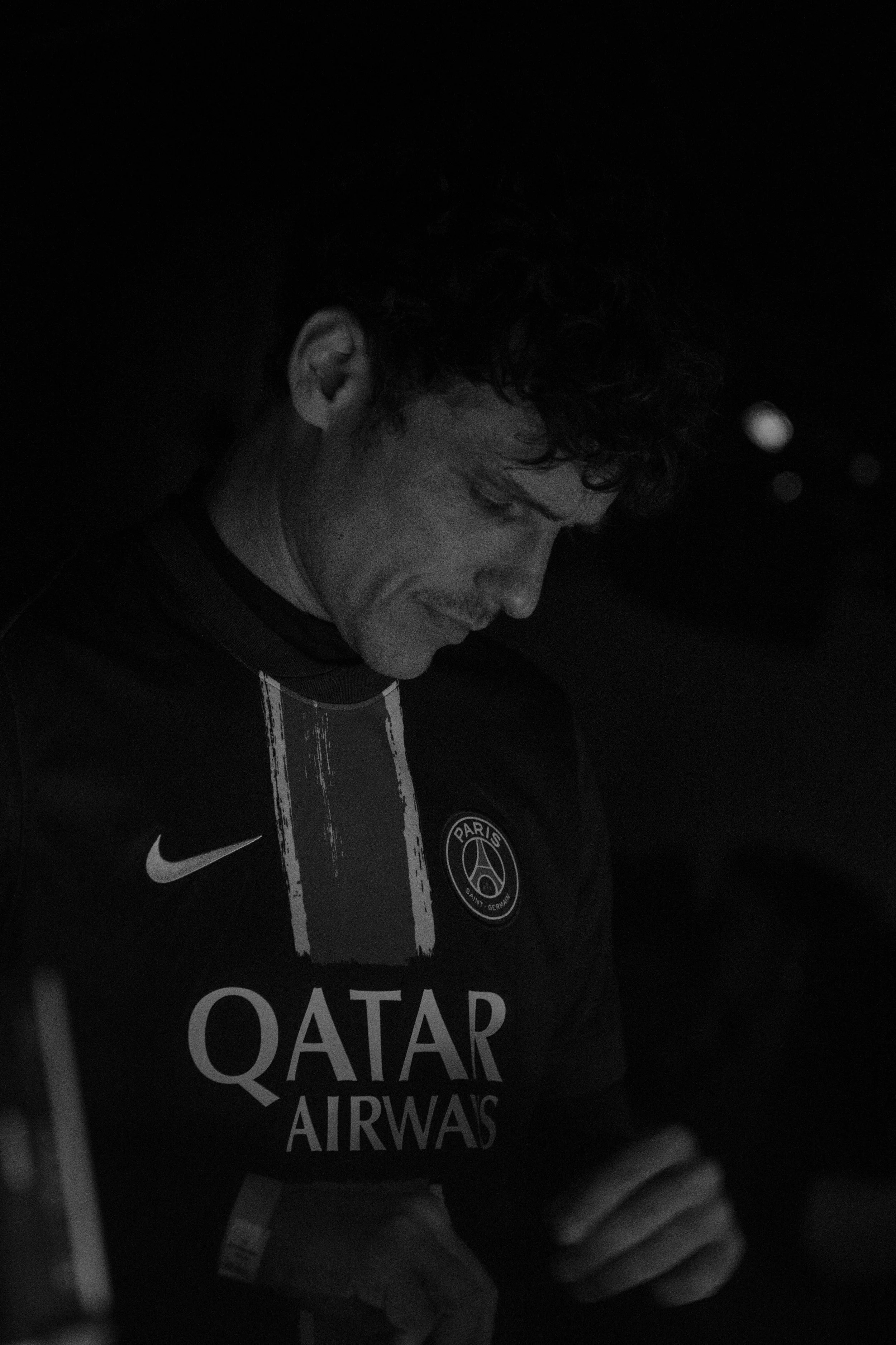 Un homme portant un maillot du Paris Saint-Germain, brandissant le logo de Qatar Airways, regarde vers le bas dans une ambiance sombre.