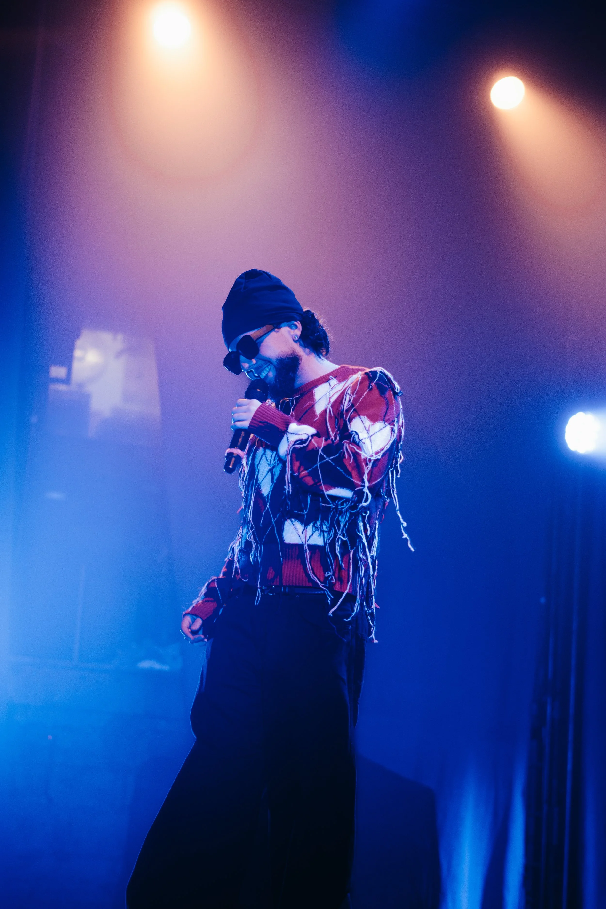 Un homme portant un chapeau noir, des lunettes de soleil, un pull rouge avec des motifs en fil blanc qui pendent, il tient un microphone en souriant sur scène avec un éclairage violet et orange.