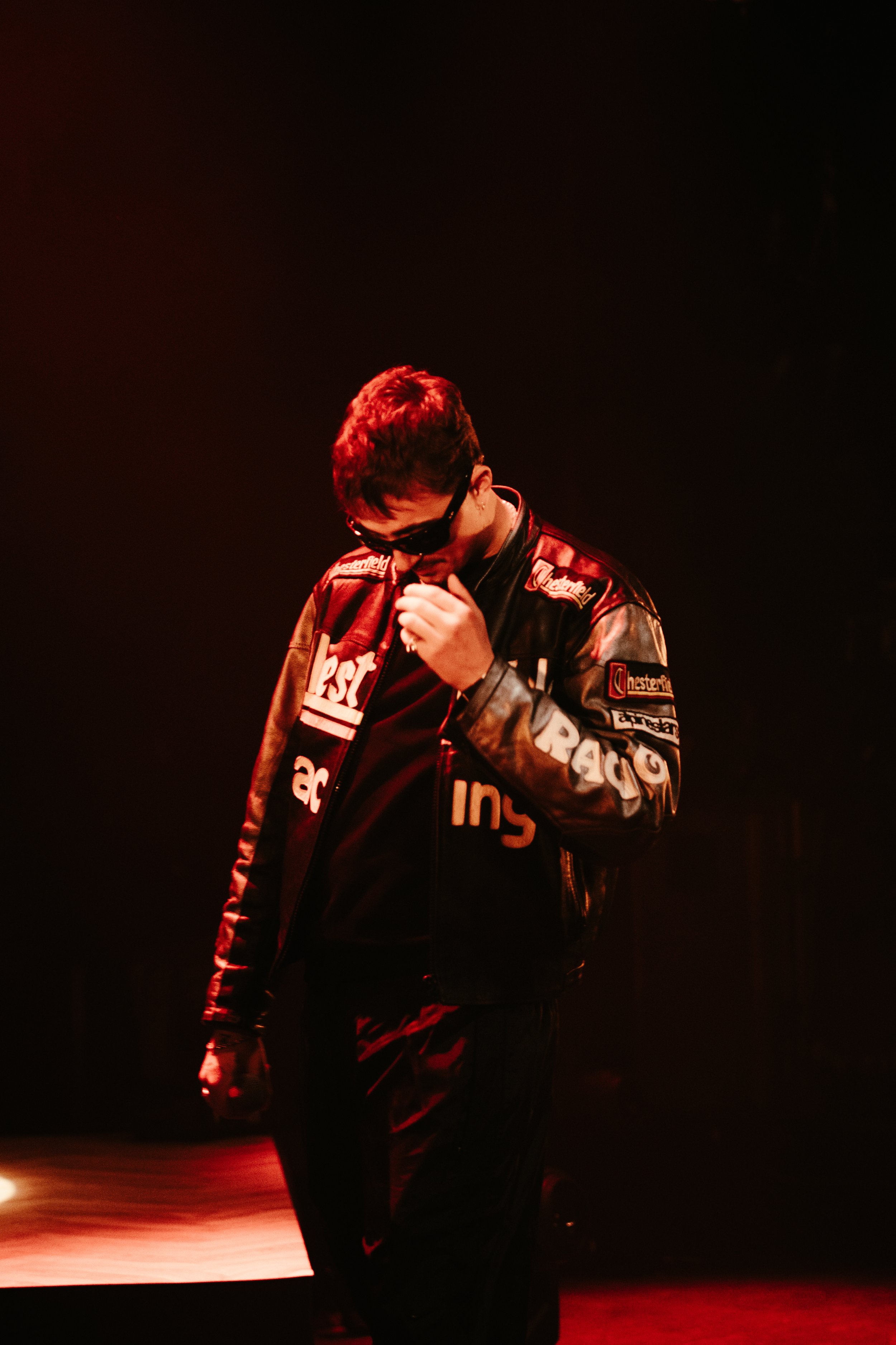 Une personne portant une veste en cuir avec des logos, portant des lunettes de soleil, dans un environnement sombre avec un éclairage rouge, semblant être en scène ou dans un studio.