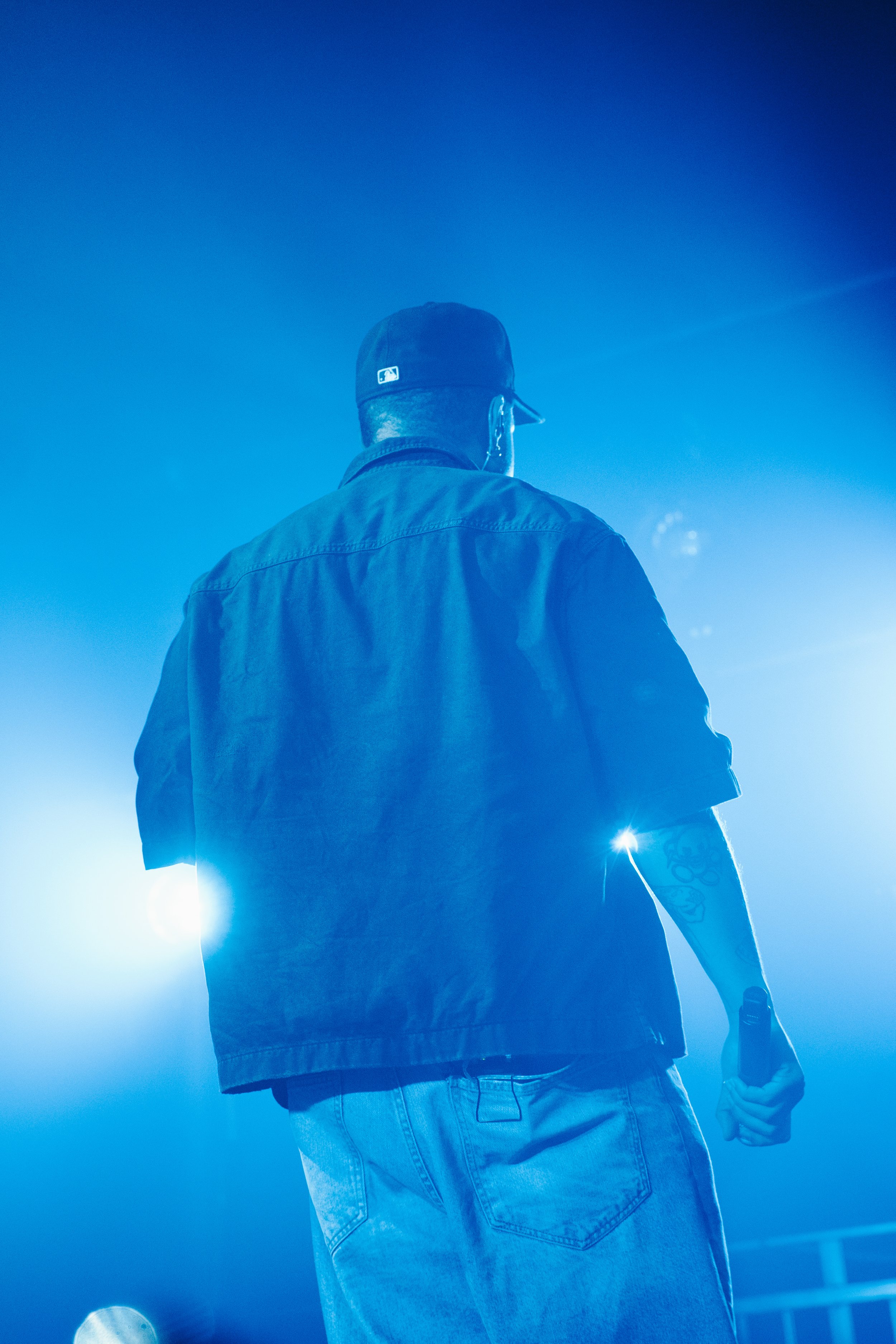 Un homme vu de dos, portant un chapeau noir, une veste et un jean, dans une ambiance de scène avec des lumières bleues.