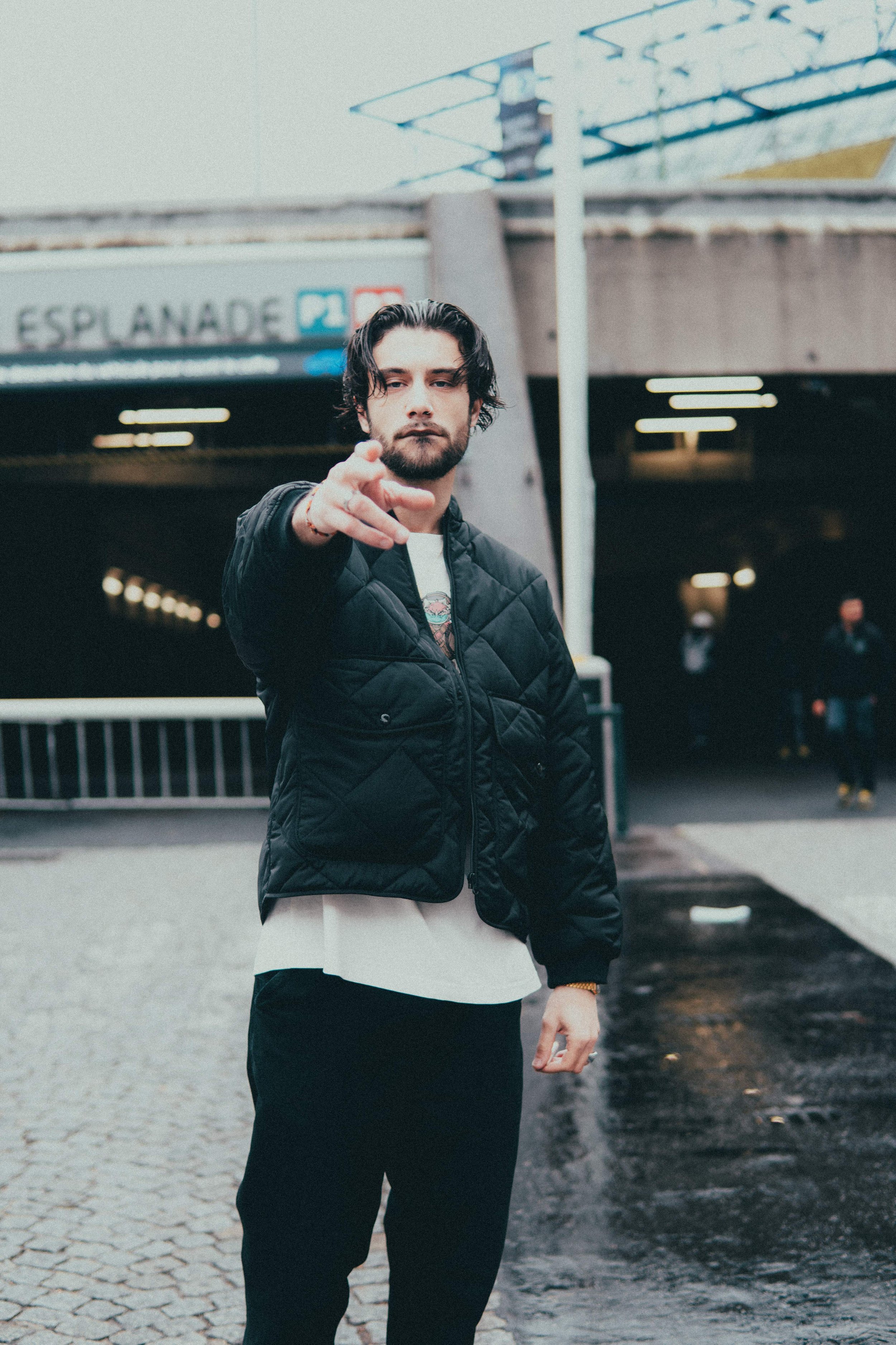 Un homme debout devant une entrée de métro, pointant vers l'appareil photo, portant une veste noire et un pantalon noir, avec un mur et un panneau "Esplanade" en arrière-plan.