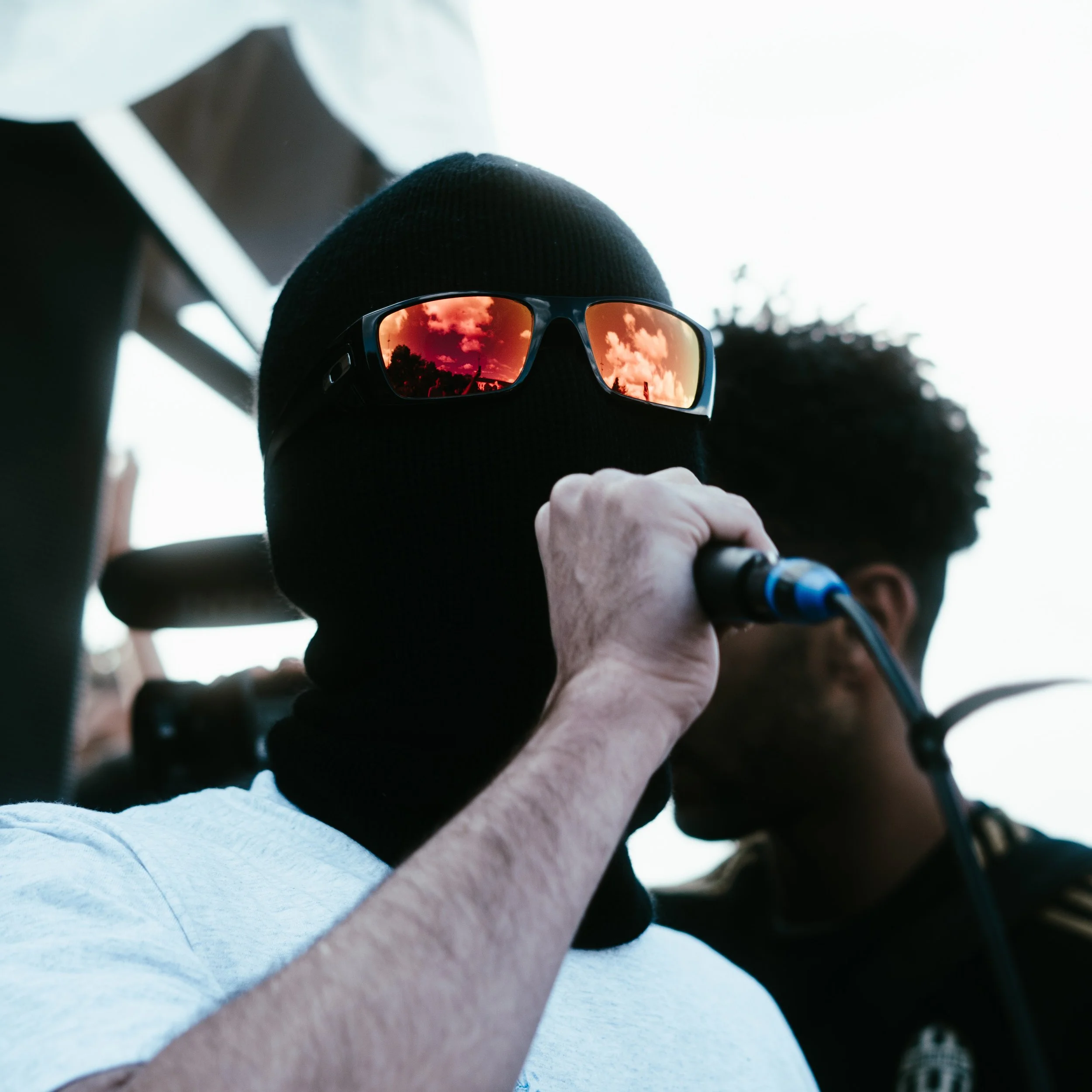 Personne portant un masque noir et des lunettes de soleil réfléchissant des nuages orangés, tenant un microphone, dans un environnement extérieur.