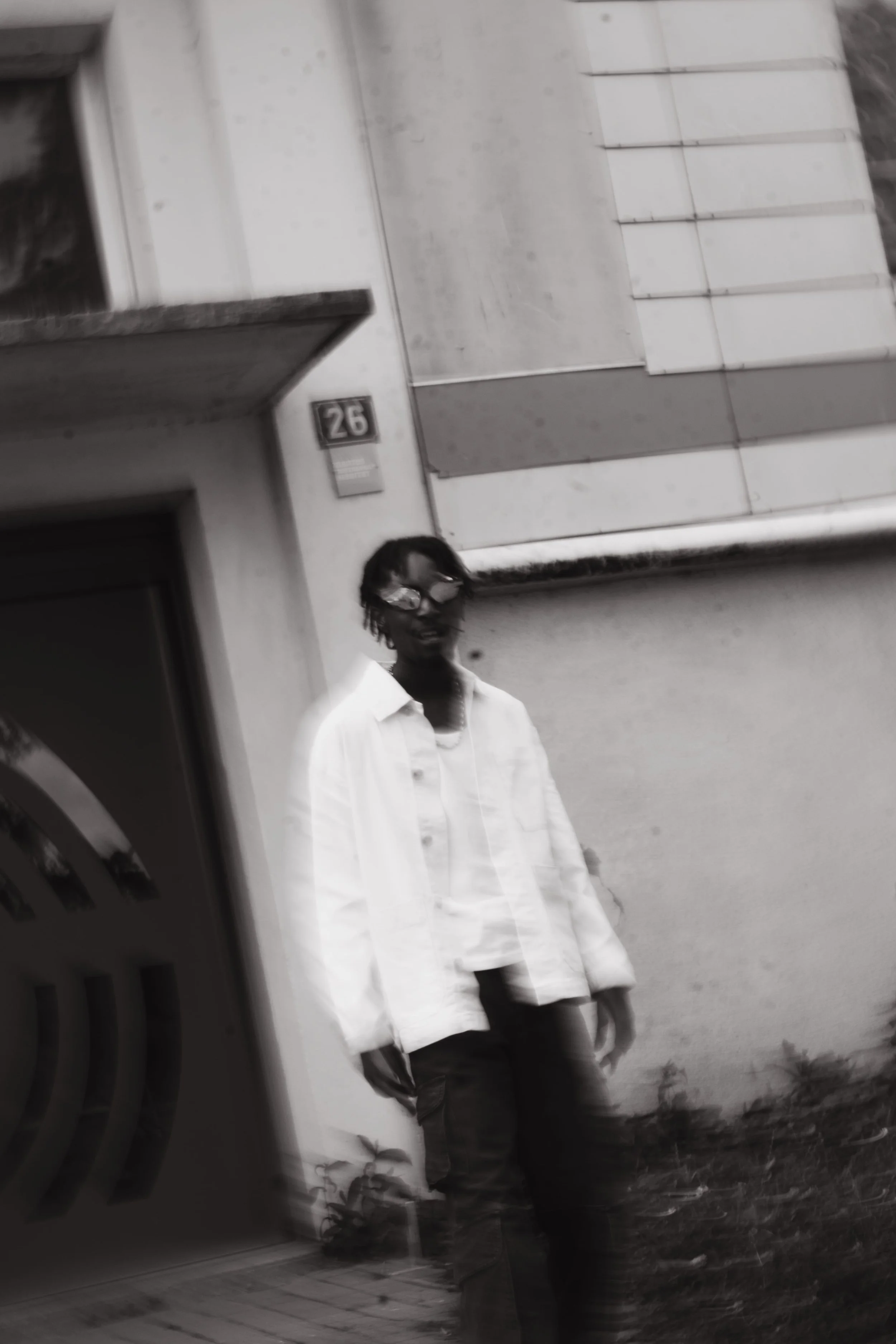 Une personne avec des lunettes, des cheveux bouclés et portant un haut blanc et un pantalon noir, marchant devant un bâtiment avec une façade en béton et une plaque portant le numéro 26.