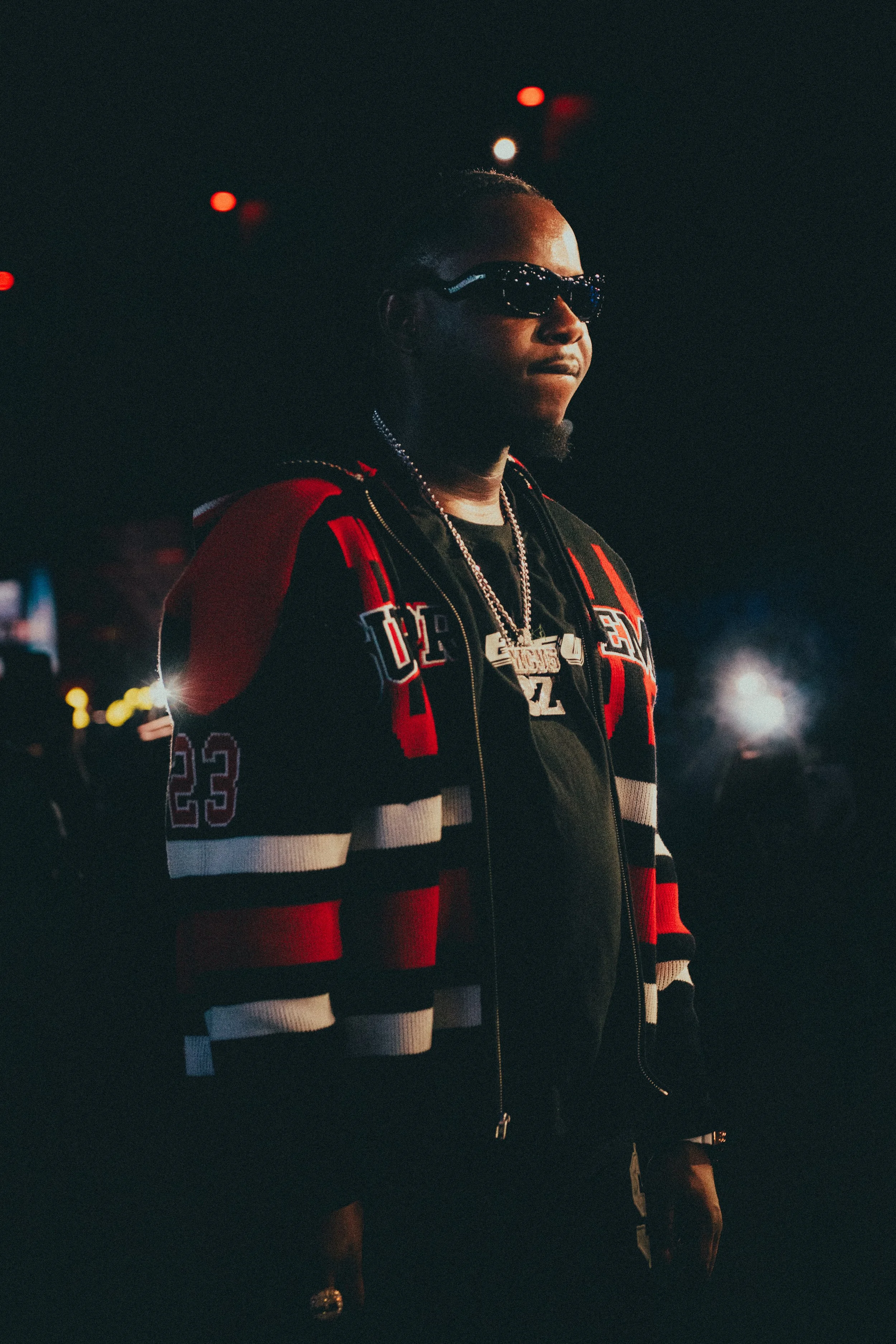 Un homme portant des lunettes de soleil, un sweat à capuche, et une veste de sport, dans un environnement sombre avec des lumières.