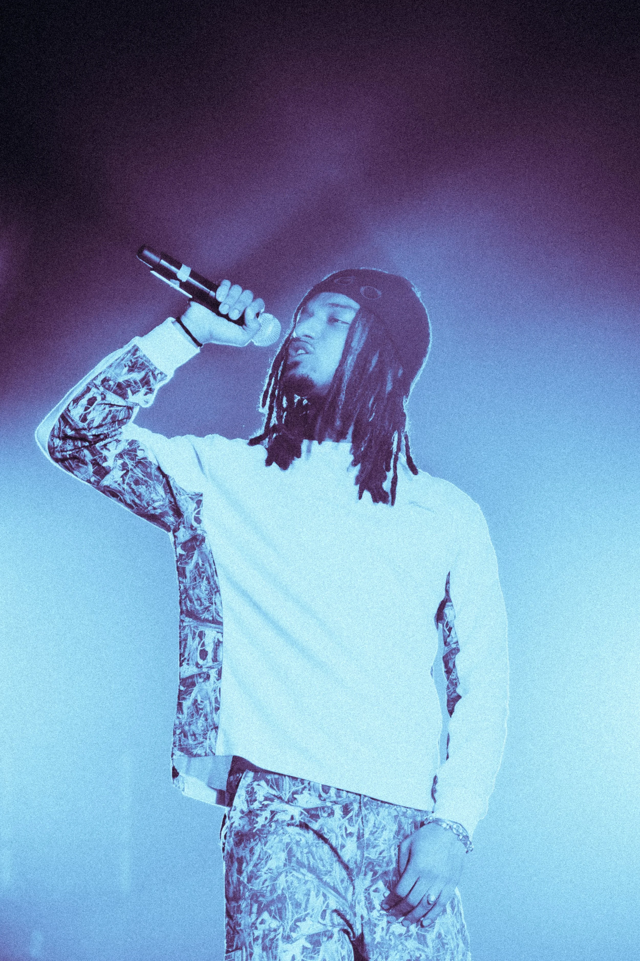 Un jeune homme avec des dreadlocks, portant un casque et un sweat à motifs, chante ou parle dans un microphone lors d'une performance, sous un éclairage bleu.