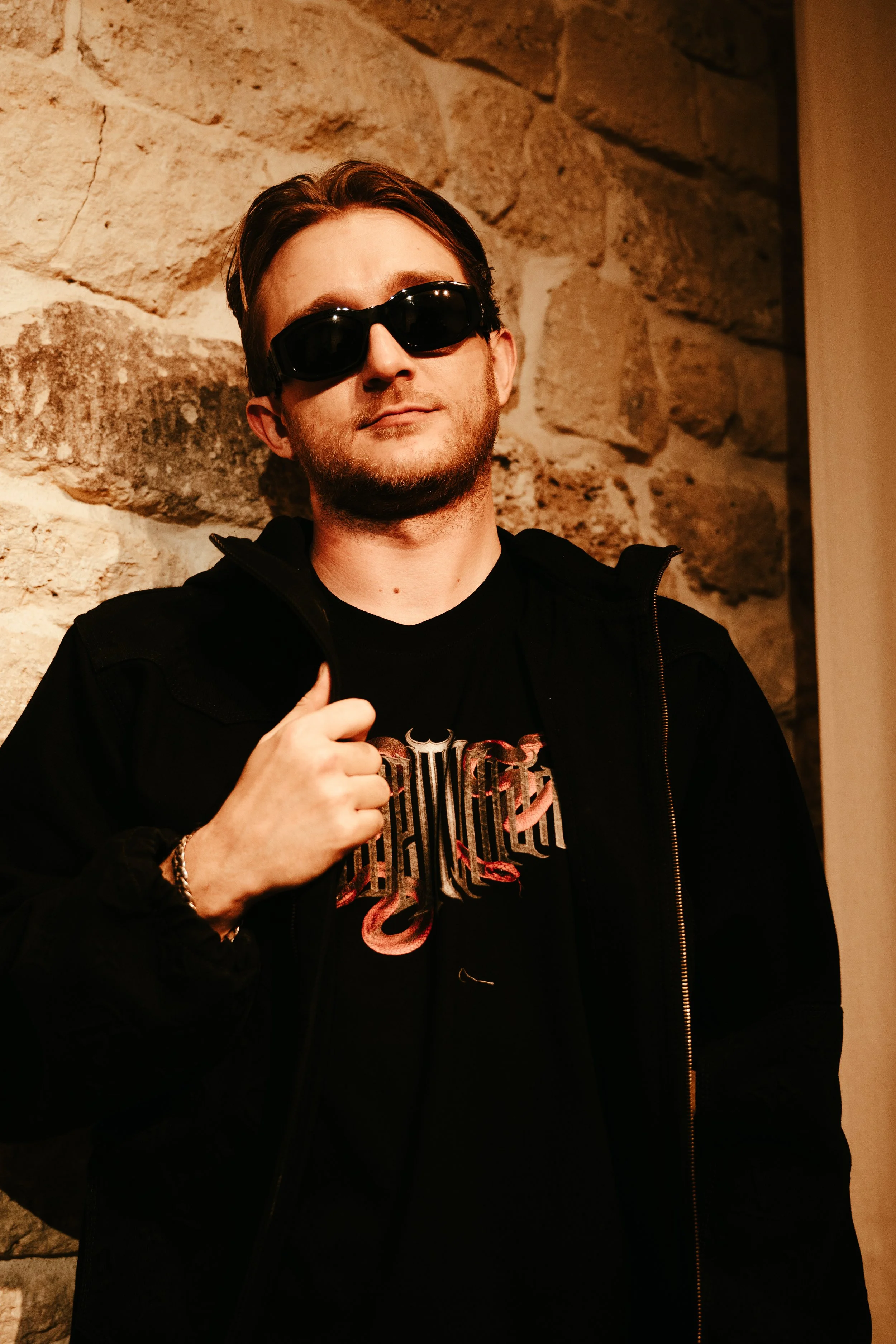 Jeune homme portant des lunettes de soleil noires, une veste noire et un t-shirt avec un design métallique, se tenant devant un mur en pierre, faisant un geste de la main.