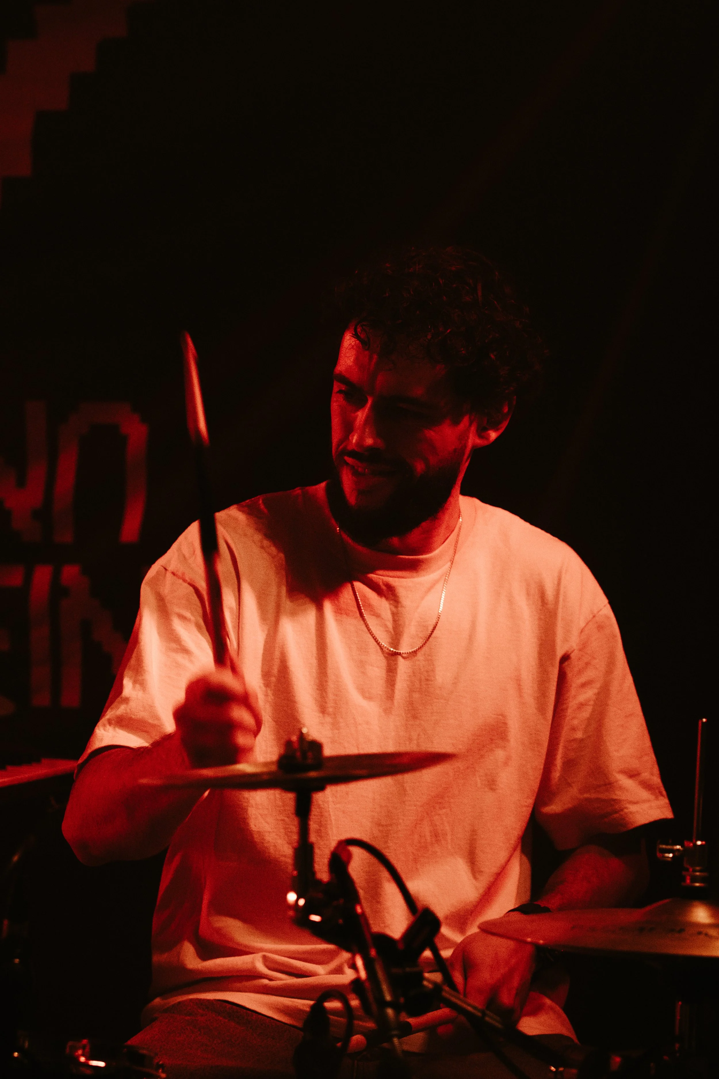 Un homme jouant de la batterie, éclairé par une lumière rouge, souriant, portant un t-shirt blanc et une chaîne en or, dans un environnement sombre.