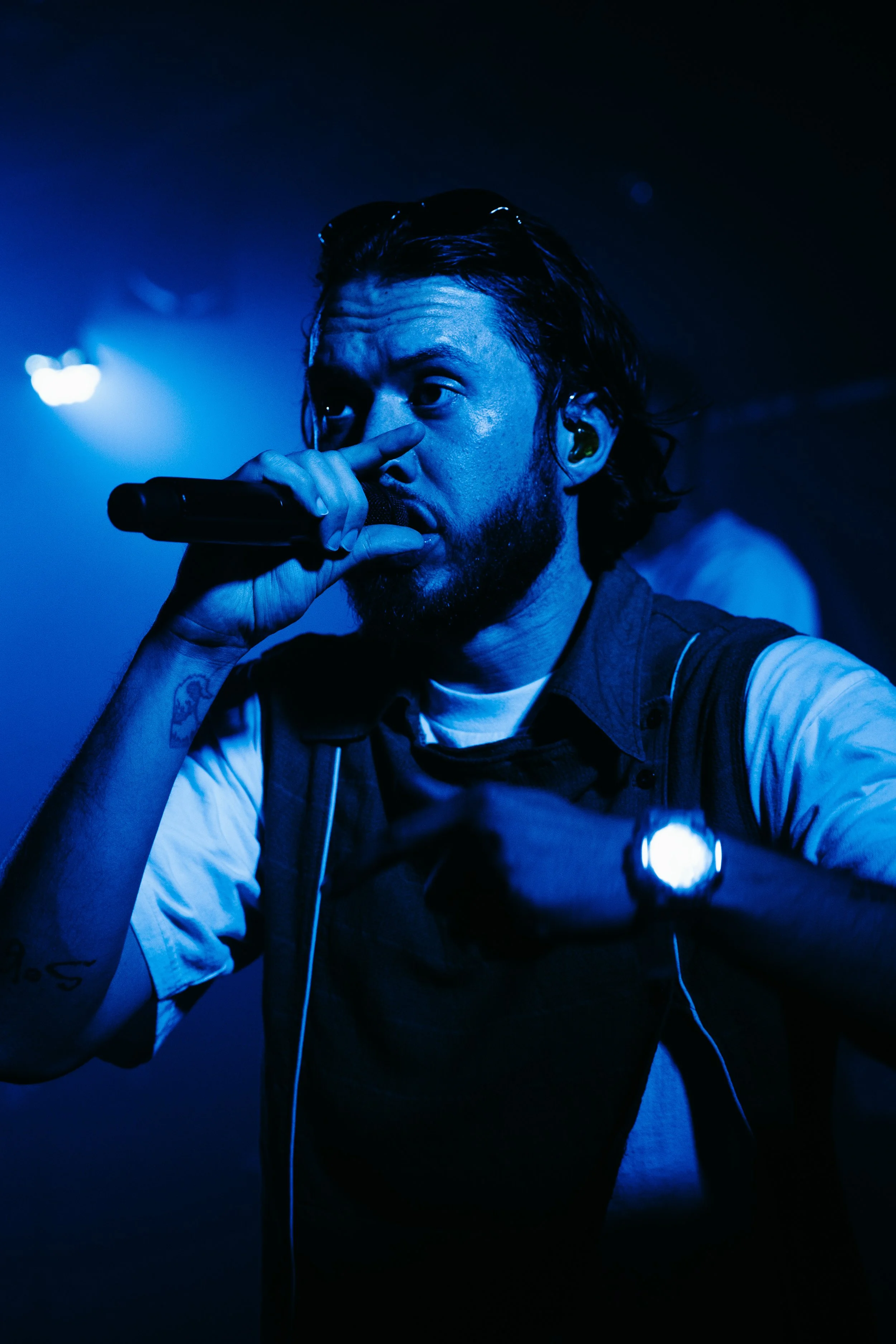 Un homme avec une barbe et des cheveux longs performe sur scène, tenant un micro, éclairé en bleu dans une ambiance de concert