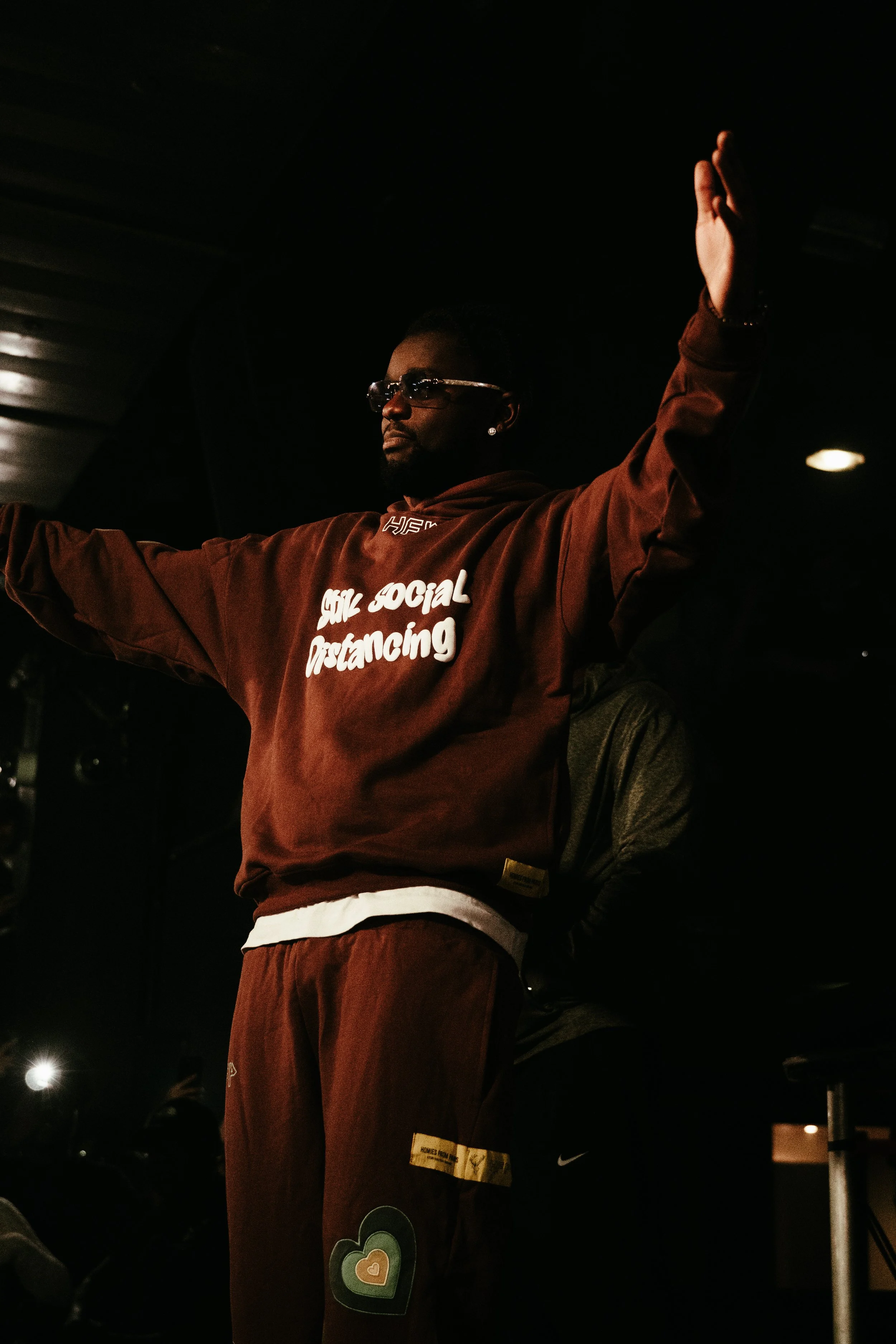 Un homme portant un sweat-shirt marron avec l'inscription "Social Distancing" en blanc, se tenant bras levés dans un environnement sombre.