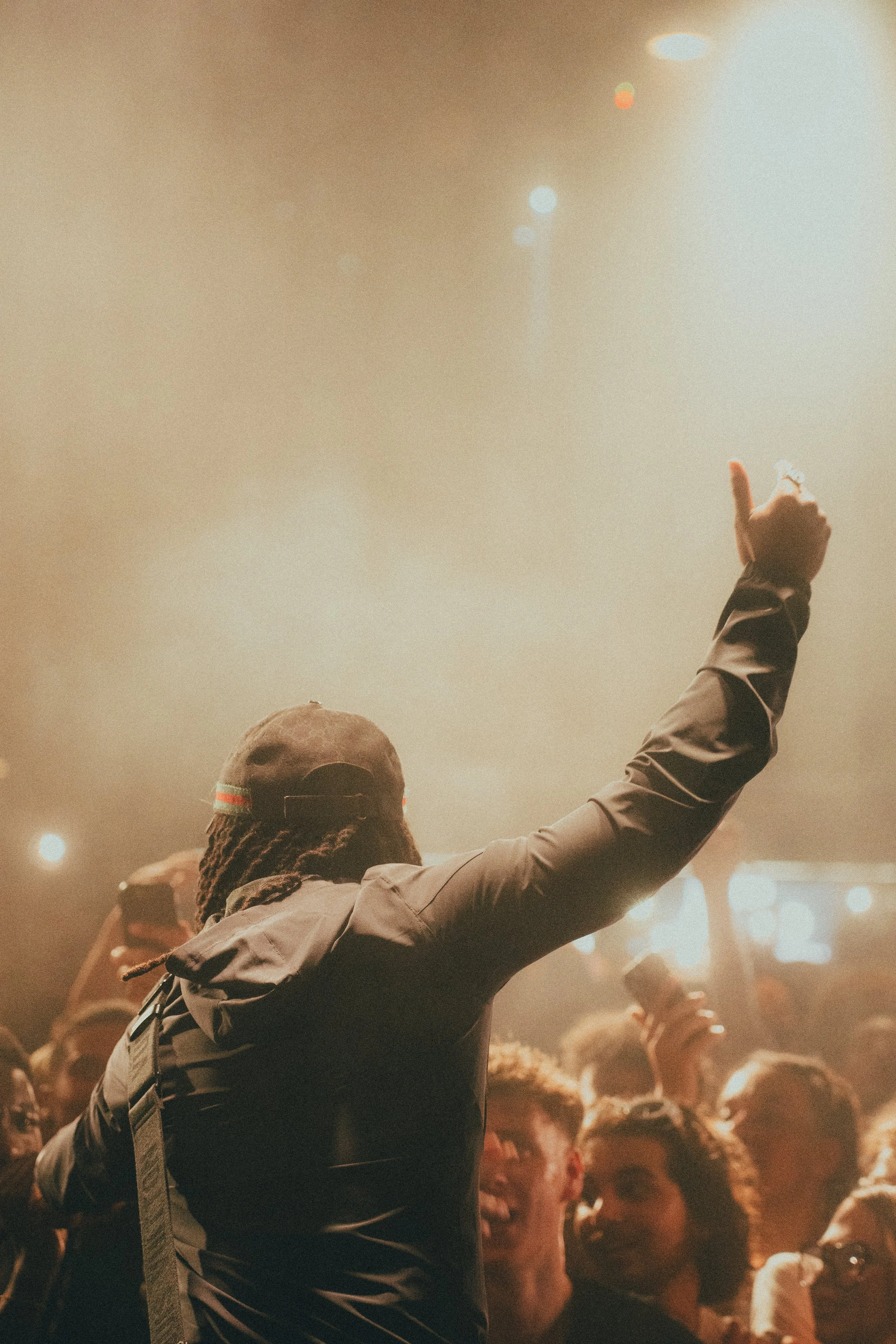 Une personne avec une casquette et une veste noire lève le bras lors d'un concert ou d'un événement avec une foule en arrière-plan.