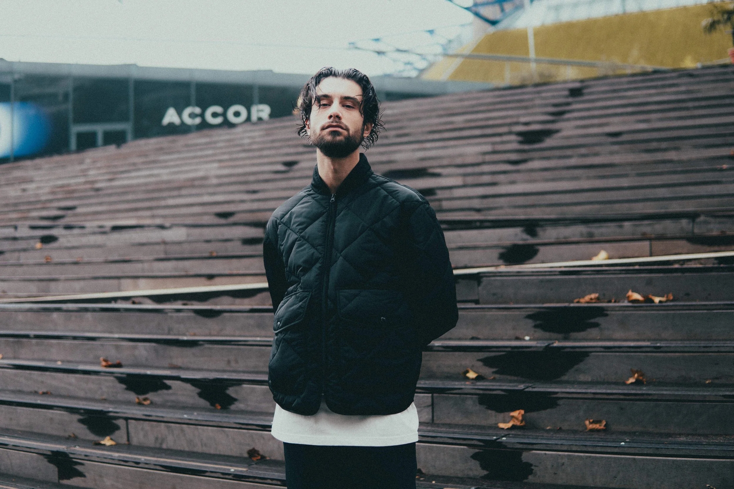 Un jeune homme avec des cheveux bouclés et une barbe, portant une veste noire, se tient devant des gradins en bois et une surface vitrée avec un panneau 'ACCOR' à l'arrière-plan.