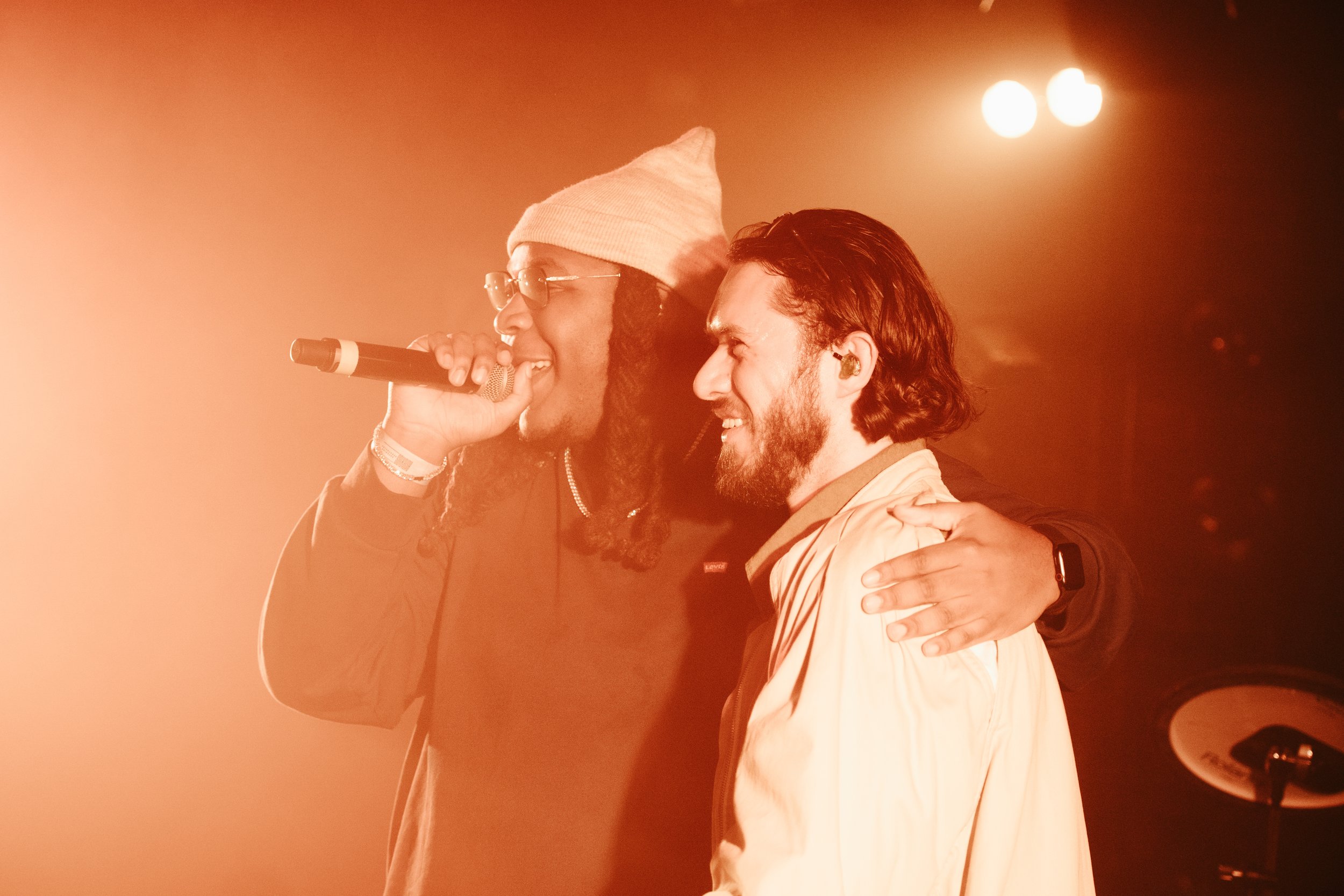 Deux hommes chantent et sourient sur scène, l'un tenant un microphone, avec un éclairage chaud et une ambiance chaleureuse.