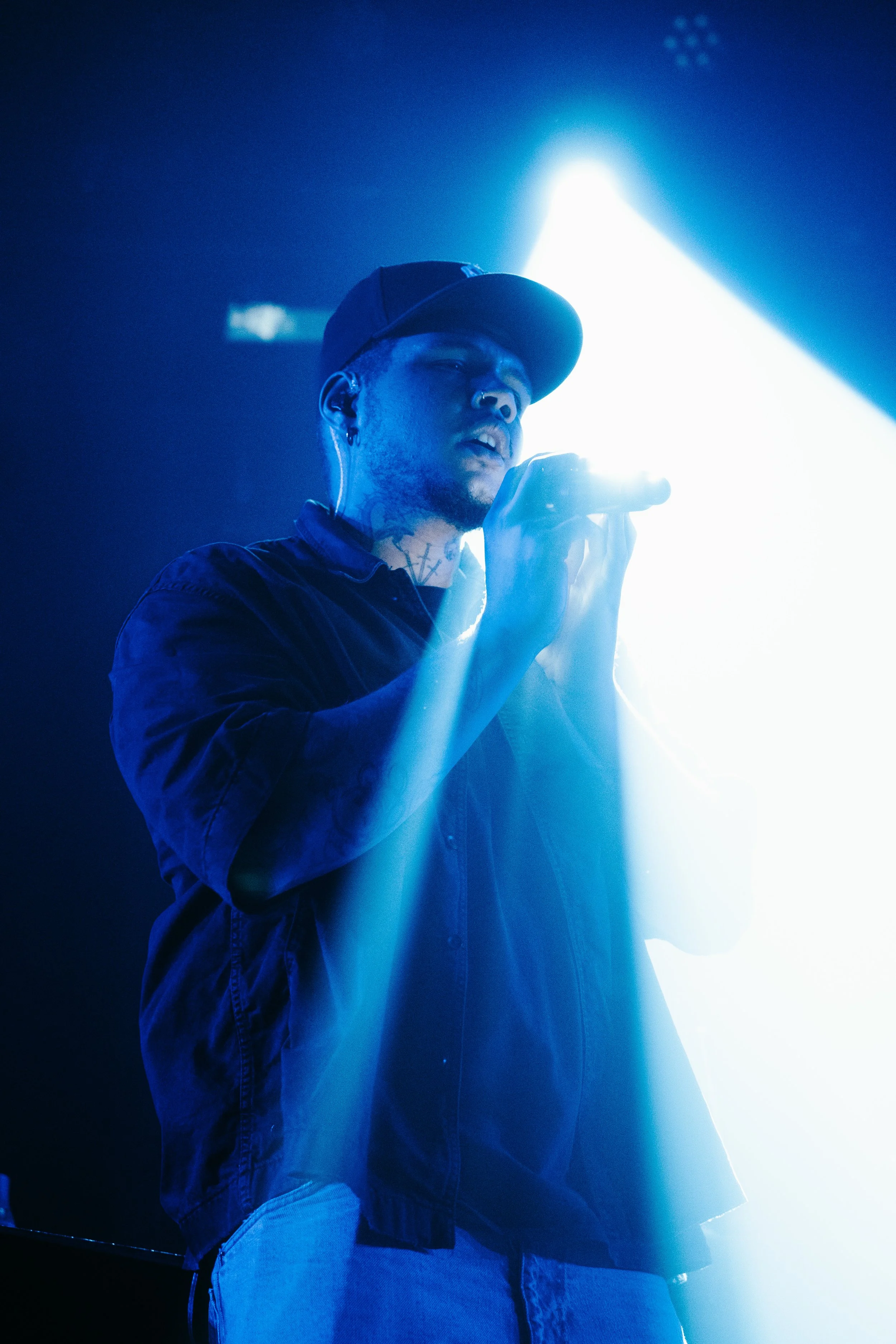 Un homme chantant ou parlant dans un micro sur scène, éclairé par une lumière bleue, portant une casquette noire, des écouteurs et une chemise noire, avec un tatouage visible sur le cou.