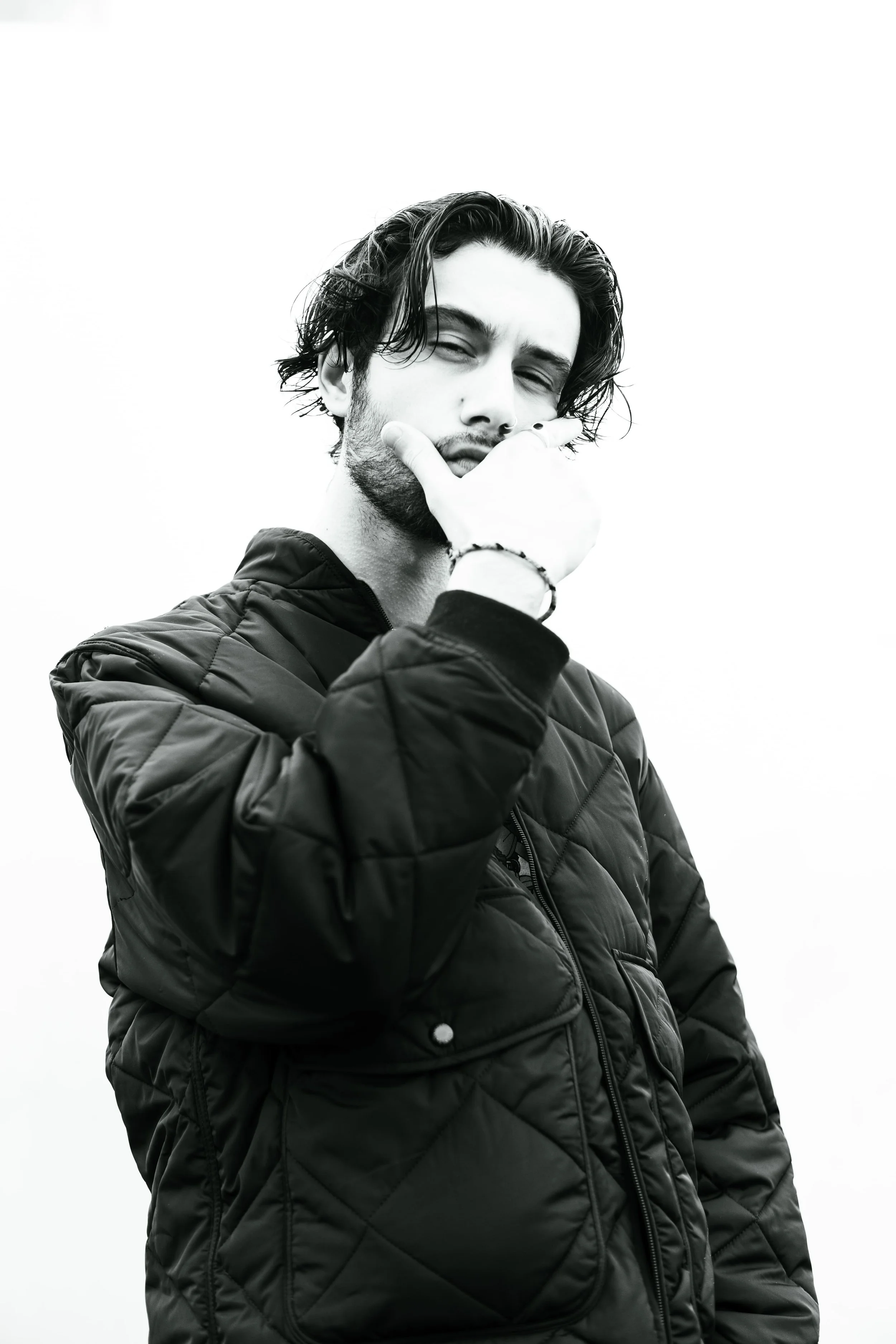 Portrait d'un jeune homme à la peau claire, avec des cheveux longs et bouclés, portant une veste rembourrée en tissu noir, se tenant contre un fond blanc, en noir et blanc.