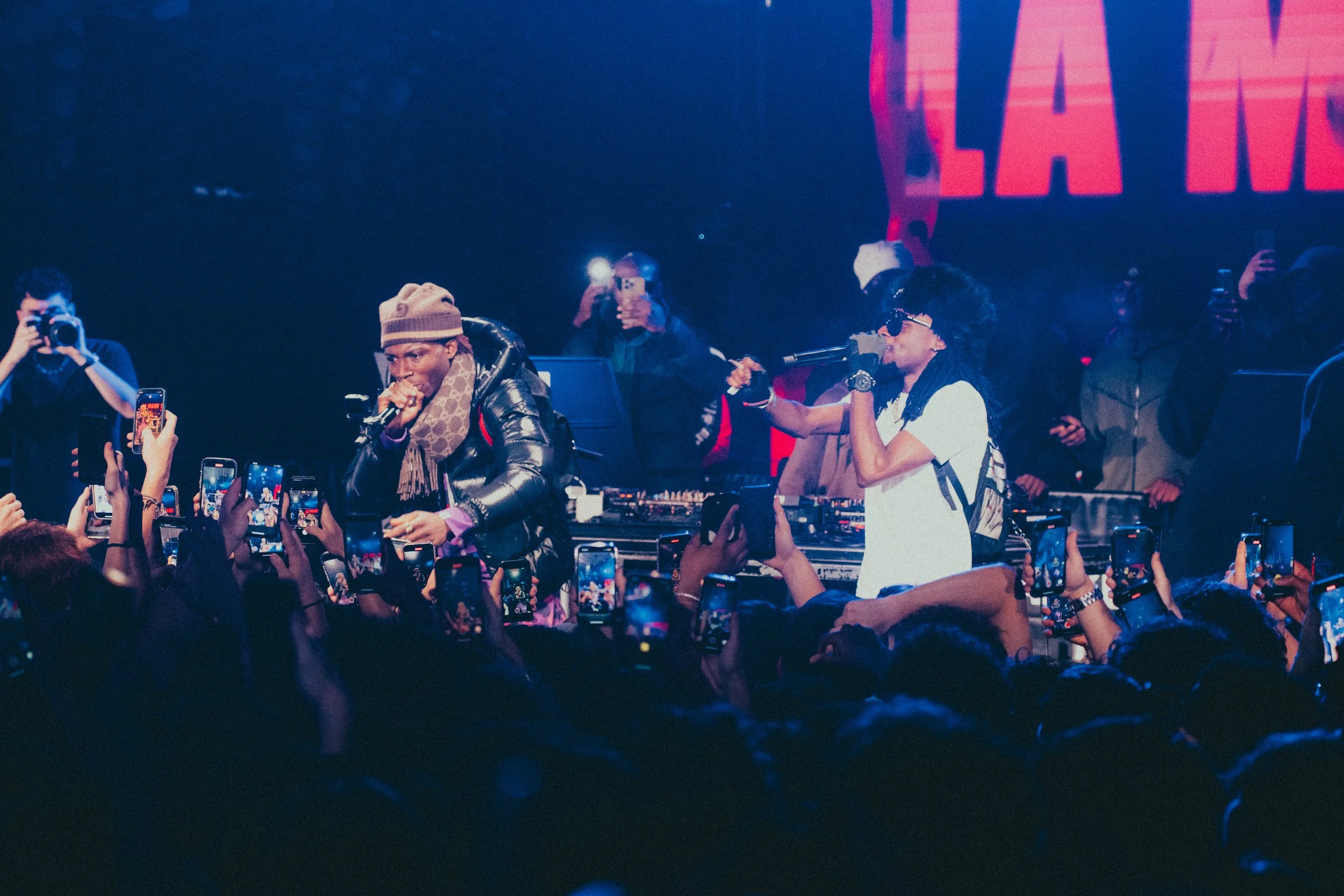 Deux artistes de rap ou de hip-hop performants sur scène lors d’un concert, entourés de nombreux spectateurs prenant des photos avec leurs téléphones.
