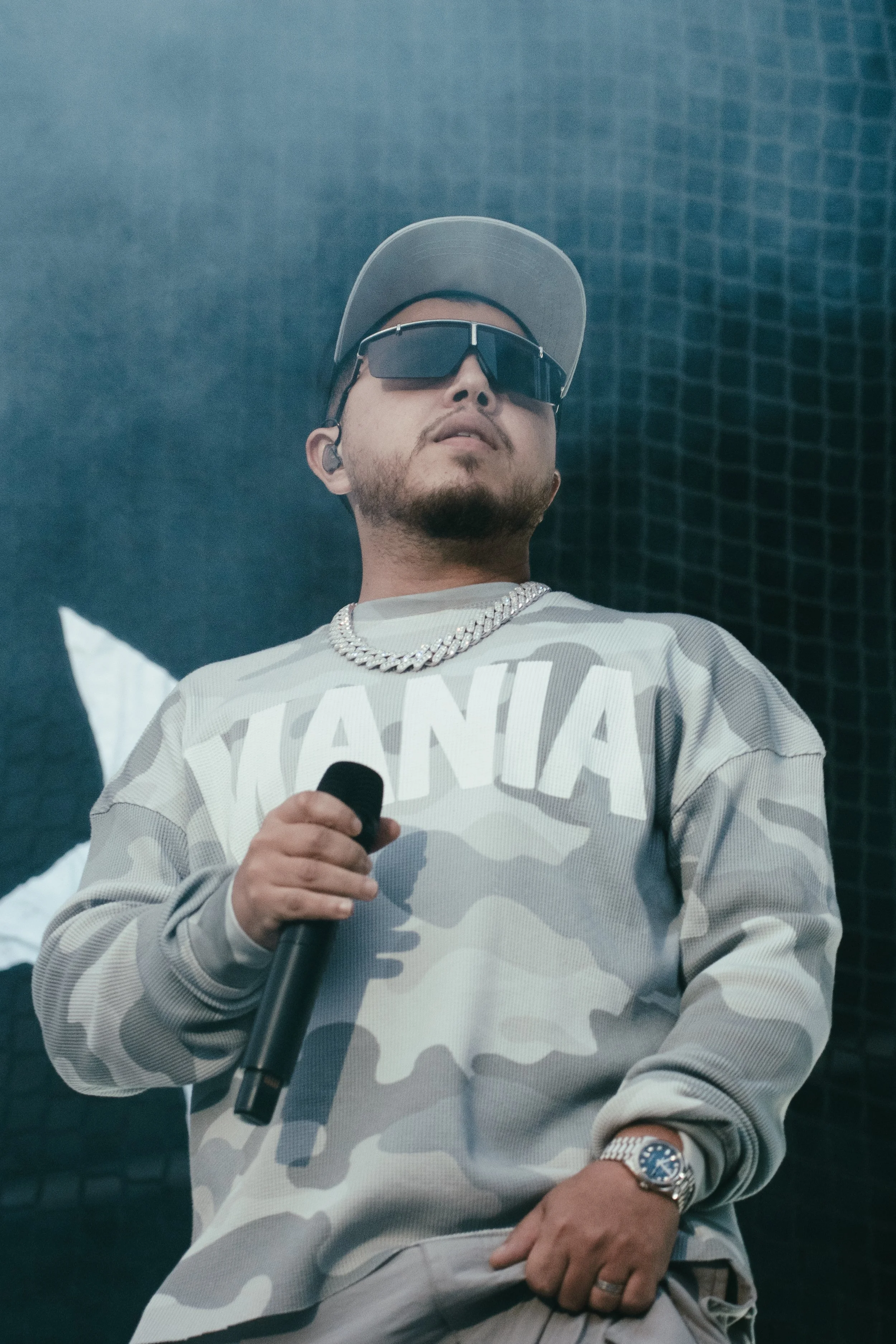 Un homme portant des lunettes de soleil, un chapeau, une chaîne en or, une montre, une veste camouflage gris avec le mot 'YAKMANIA' et tenant un microphone, sur scène.