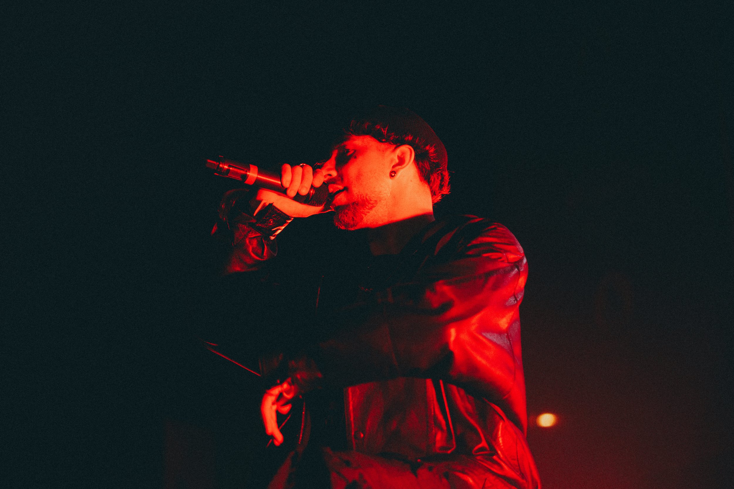 Un homme chante dans un microphone sous un éclairage rouge, portant une veste en cuir noire.