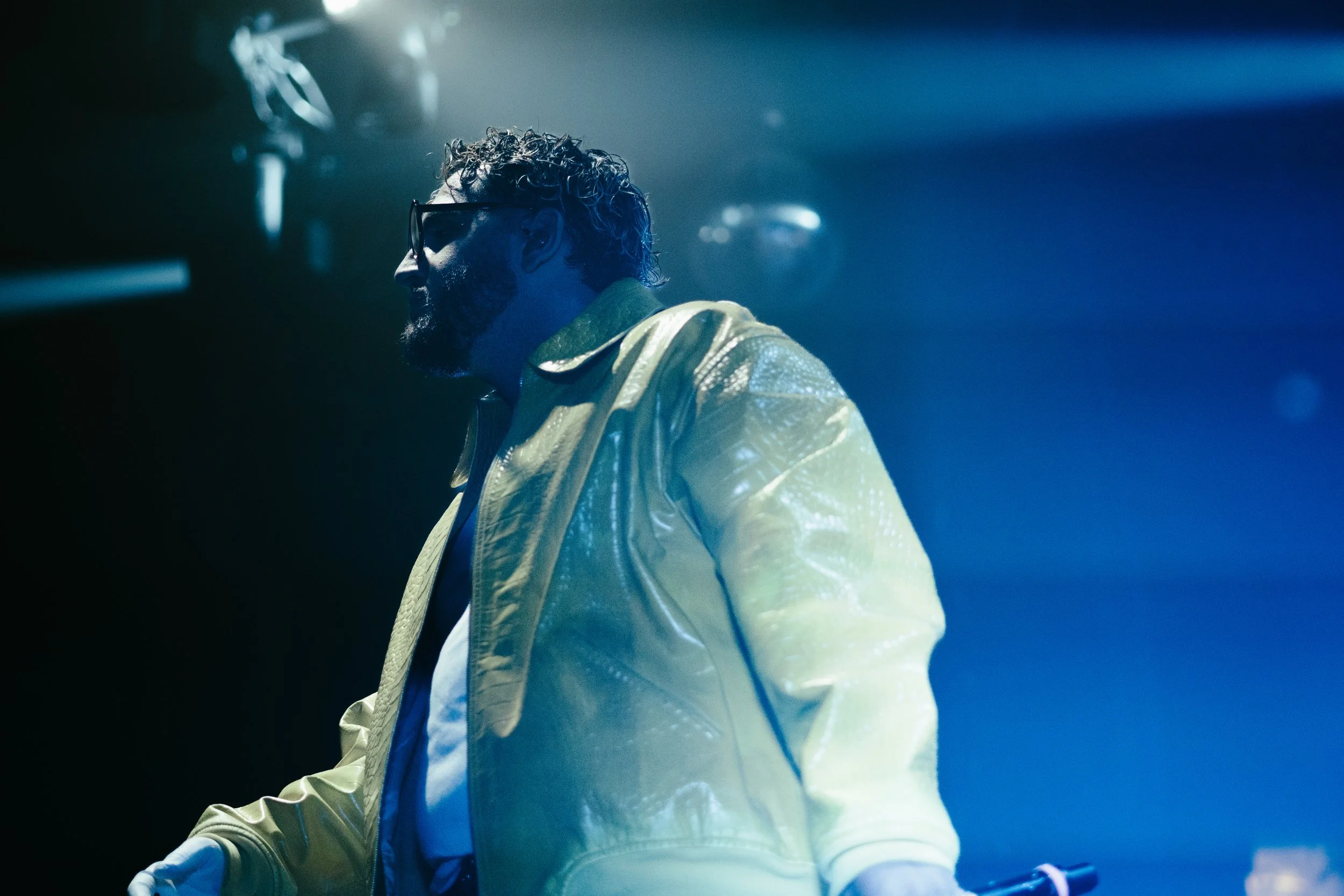 Un homme avec des cheveux bouclés, lunettes de soleil, veste jaune en cuir et barbe, dans une ambiance de scène avec lumières bleues.