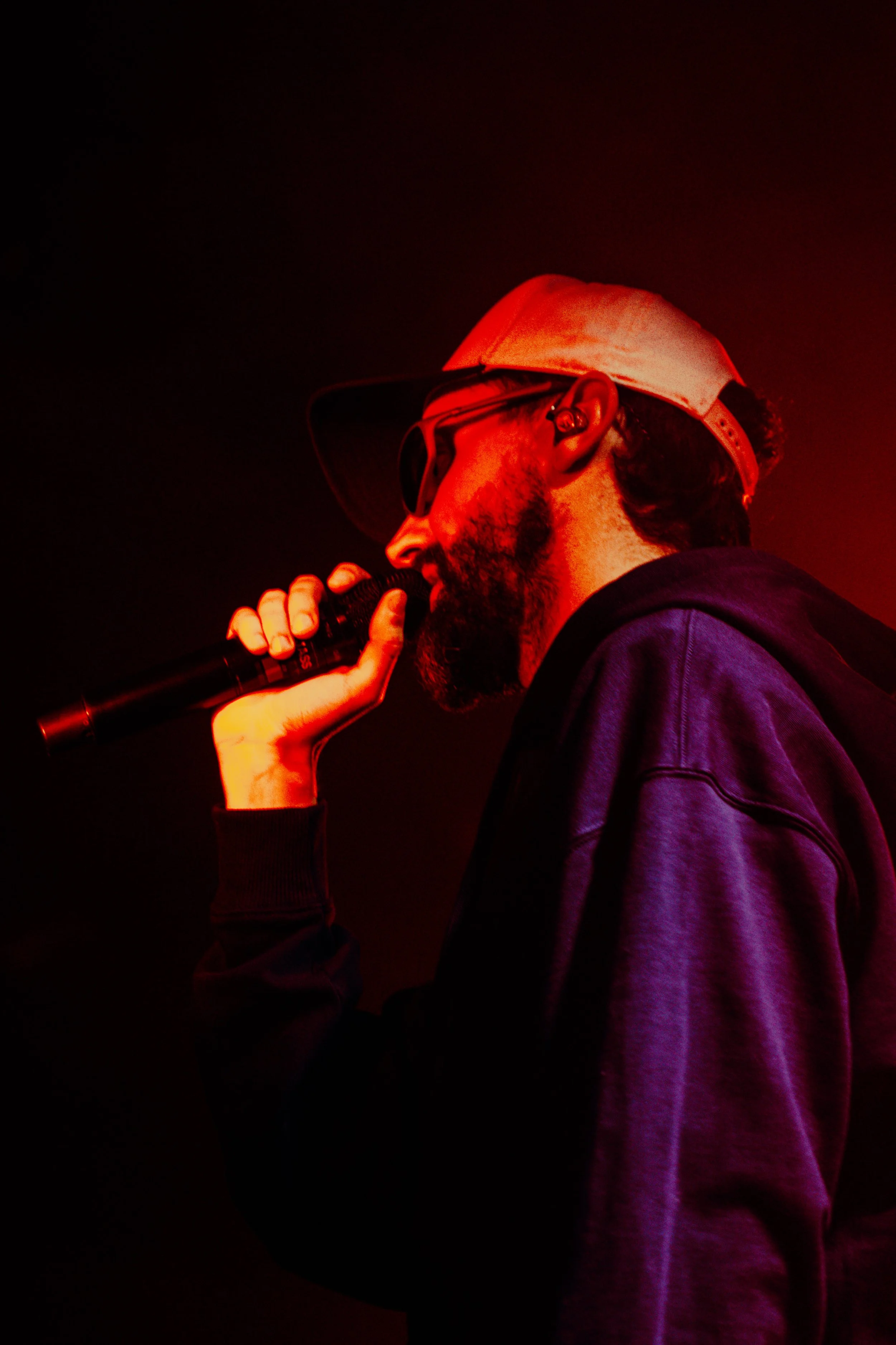 Un homme portant une casquette blanche et des lunettes de soleil noirs chante dans un microphone, avec un éclairage rouge en arrière-plan.