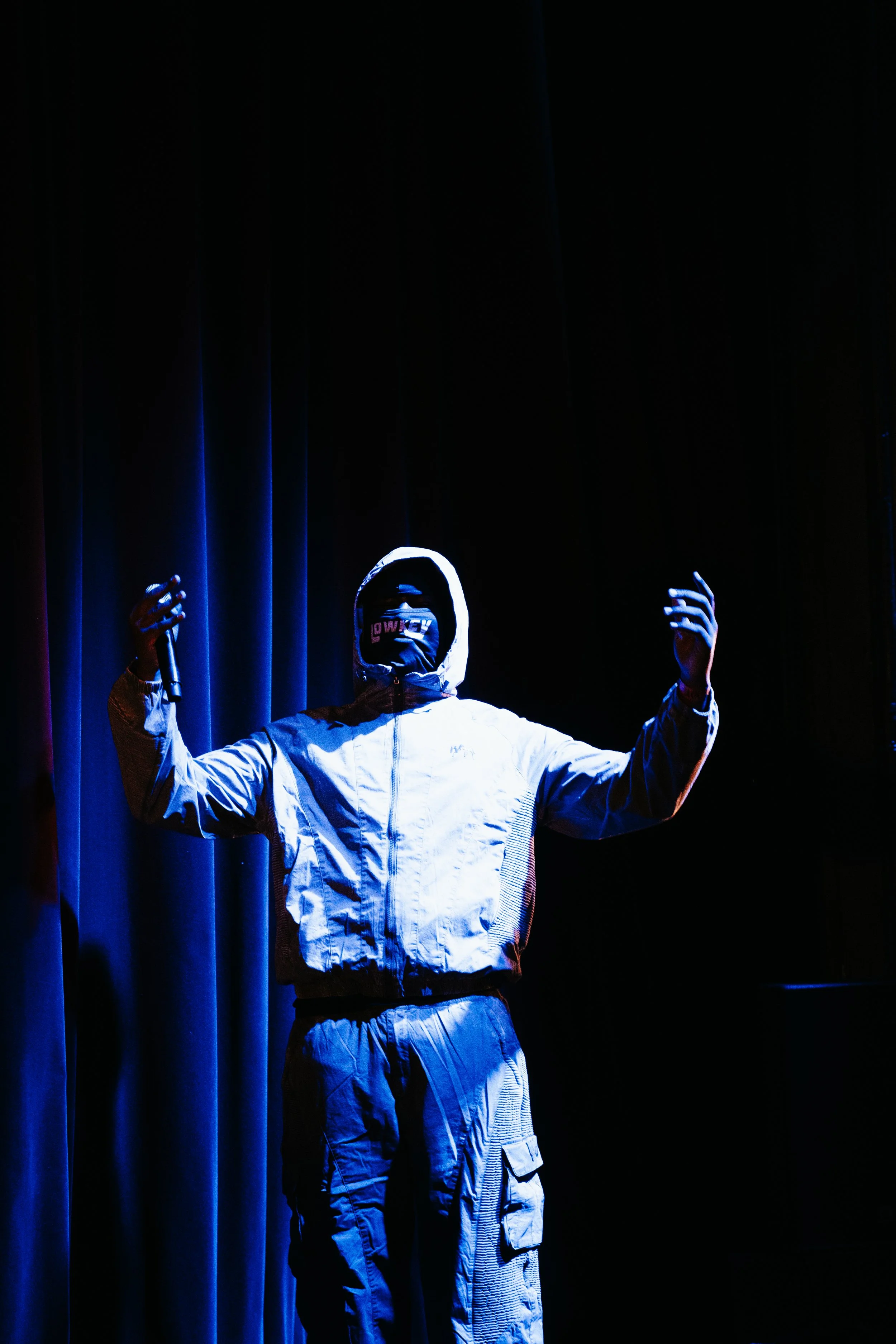 Une personne porte un sweat à capuche, un pantalon cargo, et un masque, tenant un micro, debout sur scène dans une lumière bleutée, devant un rideau noir.