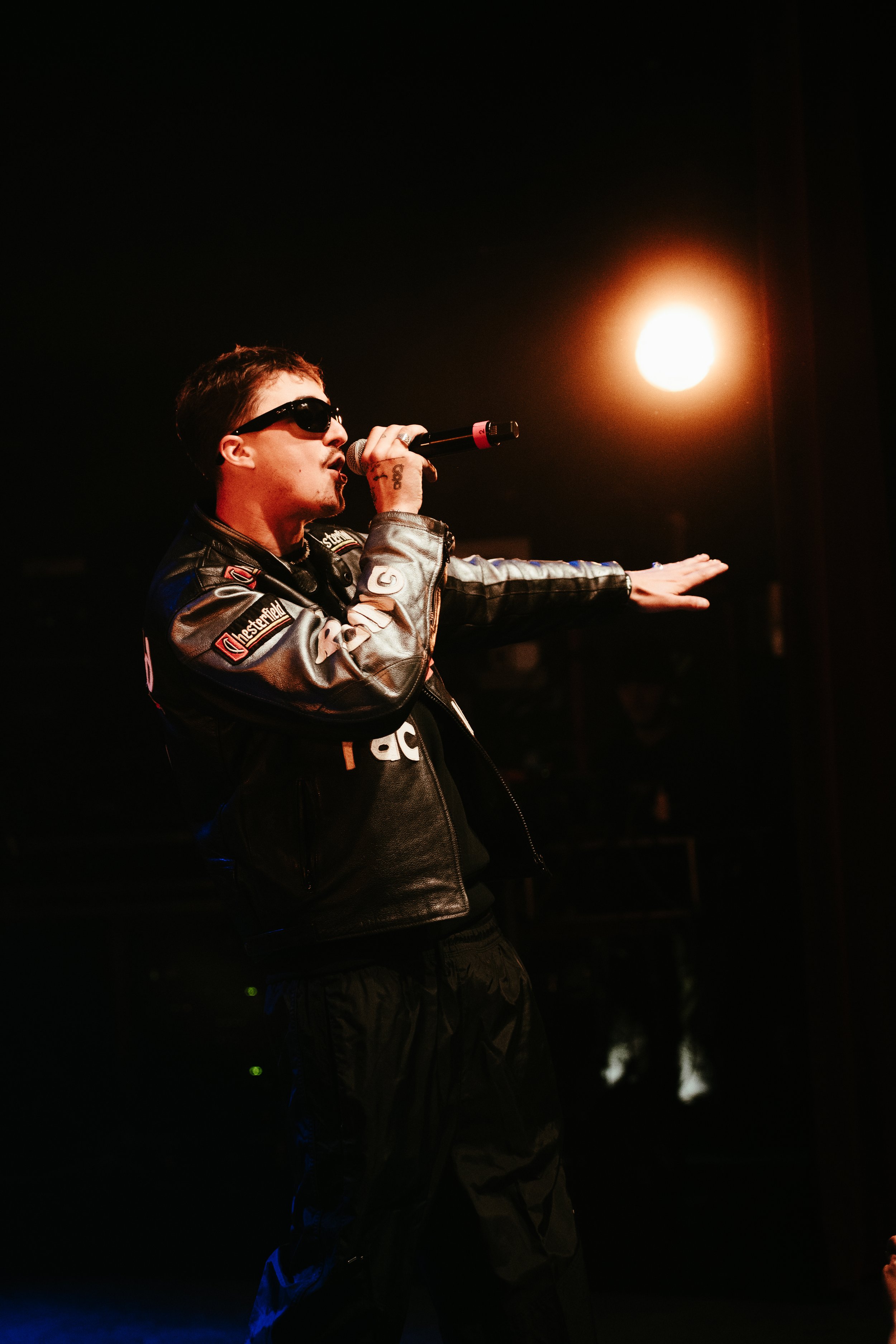 Un homme portant des lunettes de soleil noirs et une veste en cuir noire, tenant un microphone, en train de performer sur scène avec un éclairage chaud derrière lui.