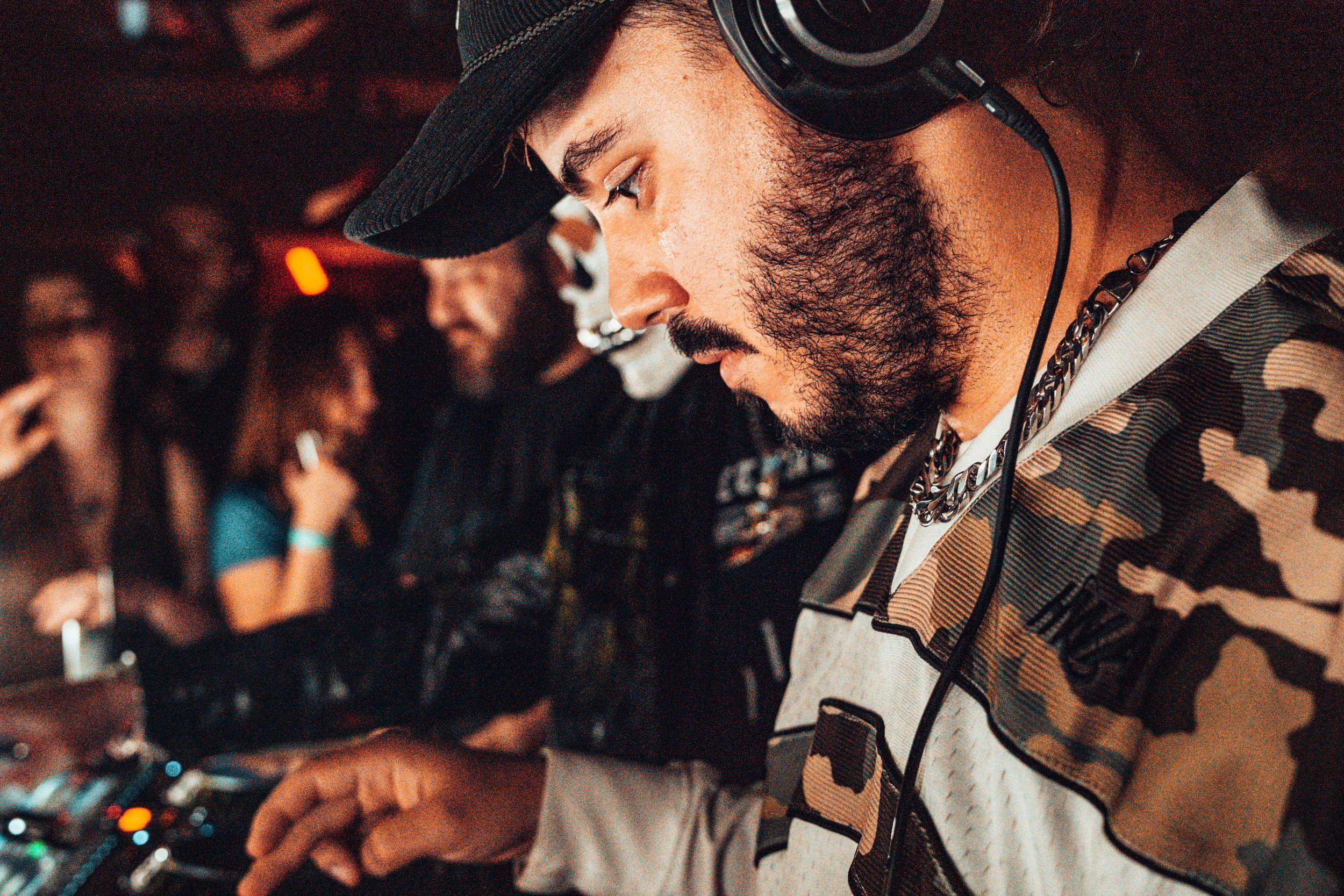 DJ avec casque, portant un sweat à capuche camouflage, en train de mixer dans une ambiance de fête, avec un groupe de personnes en arrière-plan.