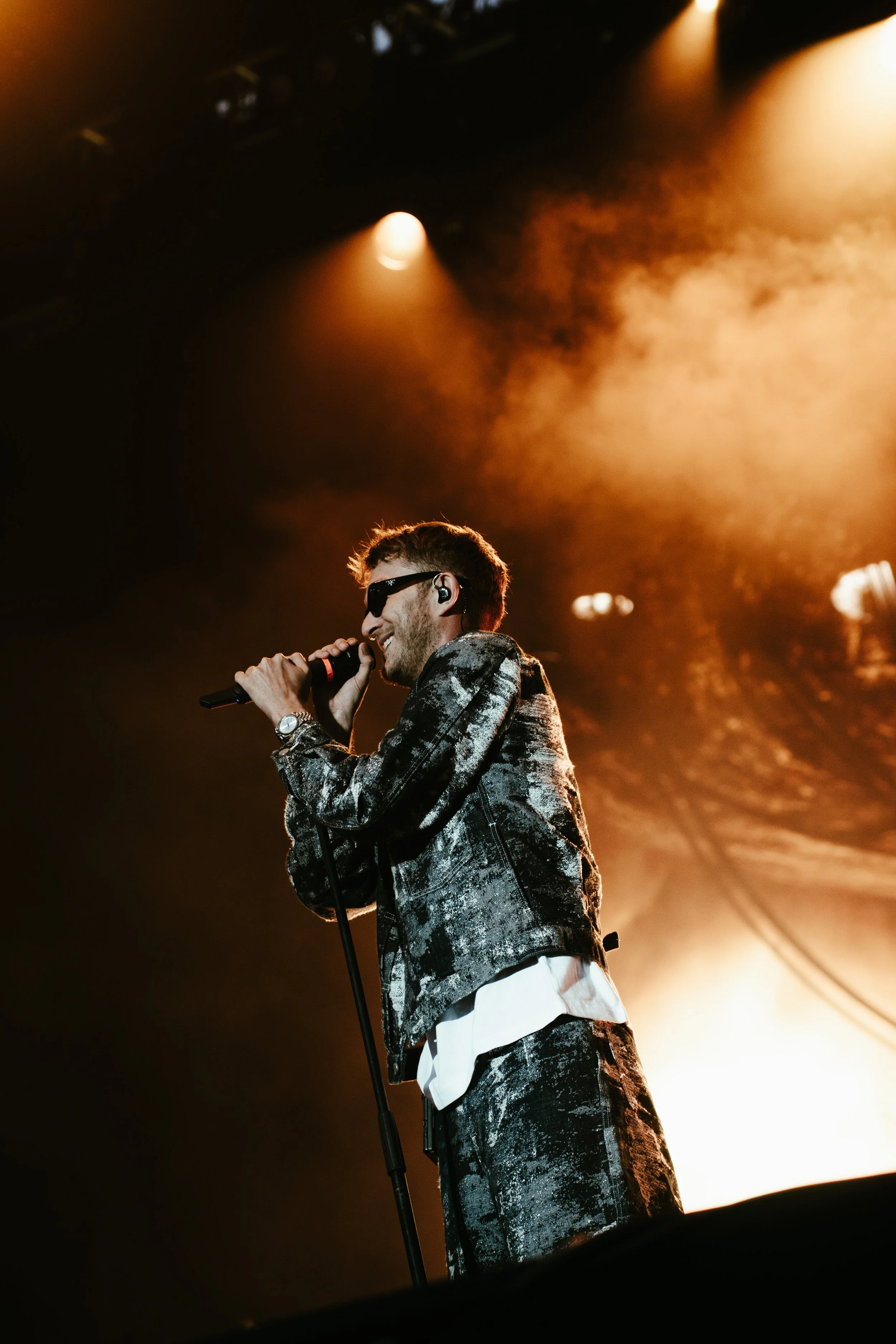 Un chanteur sur scène, portant des lunettes de soleil, une veste noire et une chemise blanche, tenant un microphone lumineux, sous un éclairage chaud et brumeux.