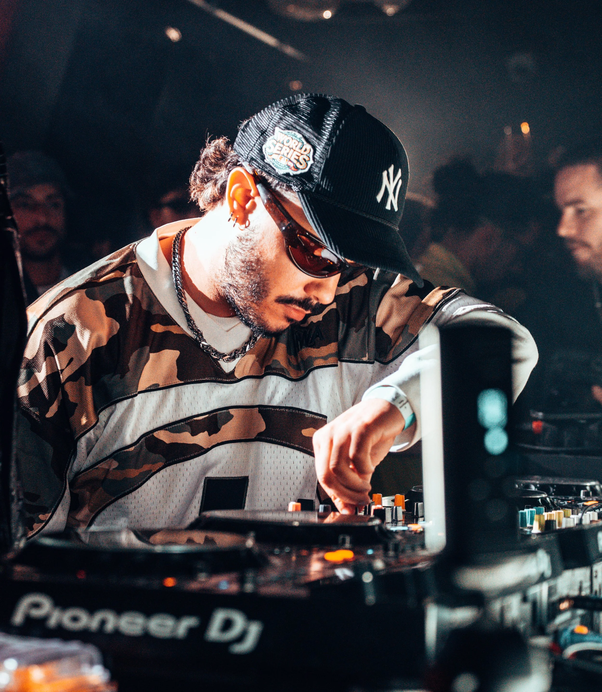 DJ jouant dans une boîte de nuit, portant une casquette New York Yankees, des lunettes de soleil, un t-shirt camouflage et un collier.