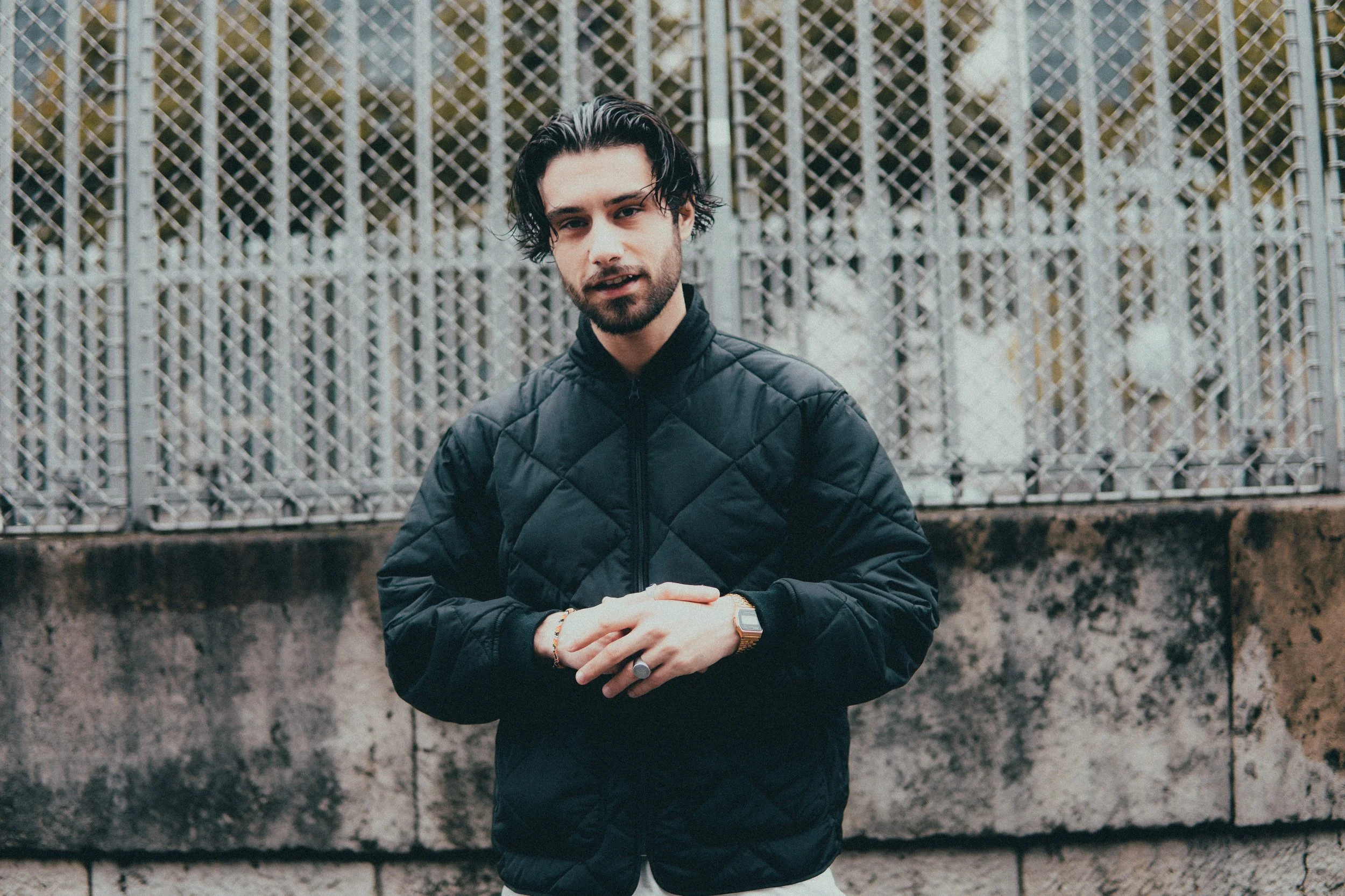 Jeune homme avec cheveux noirs, barbe, portant une veste noire, debout devant une grille en métal et un mur en pierre, regardant vers la caméra.