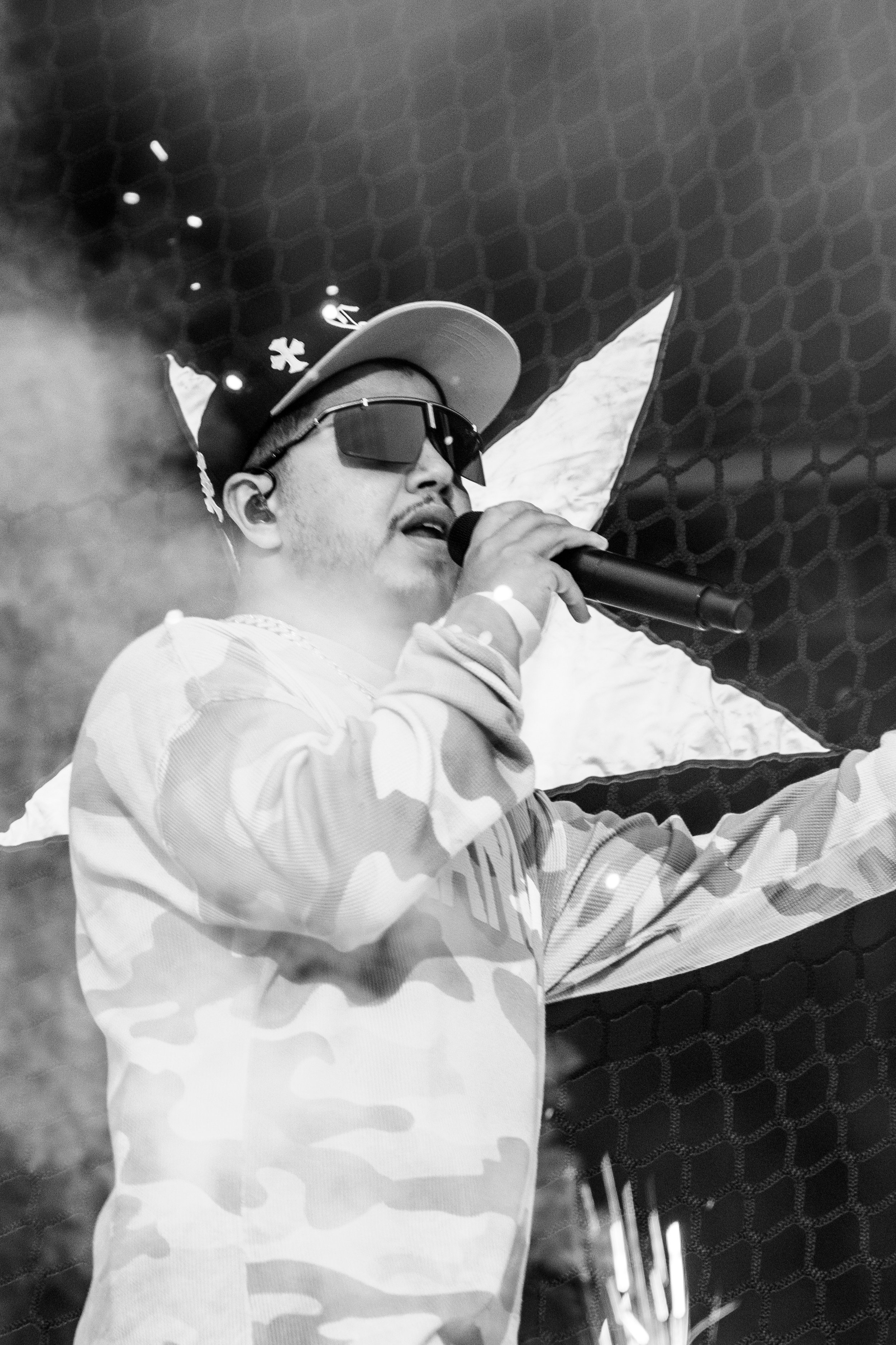 Un homme portant des lunettes de soleil, un chapeau, des écouteurs, un sweat à capuche camouflage, tenant un micro et chantant ou parlant, avec un drapeau en arrière-plan.