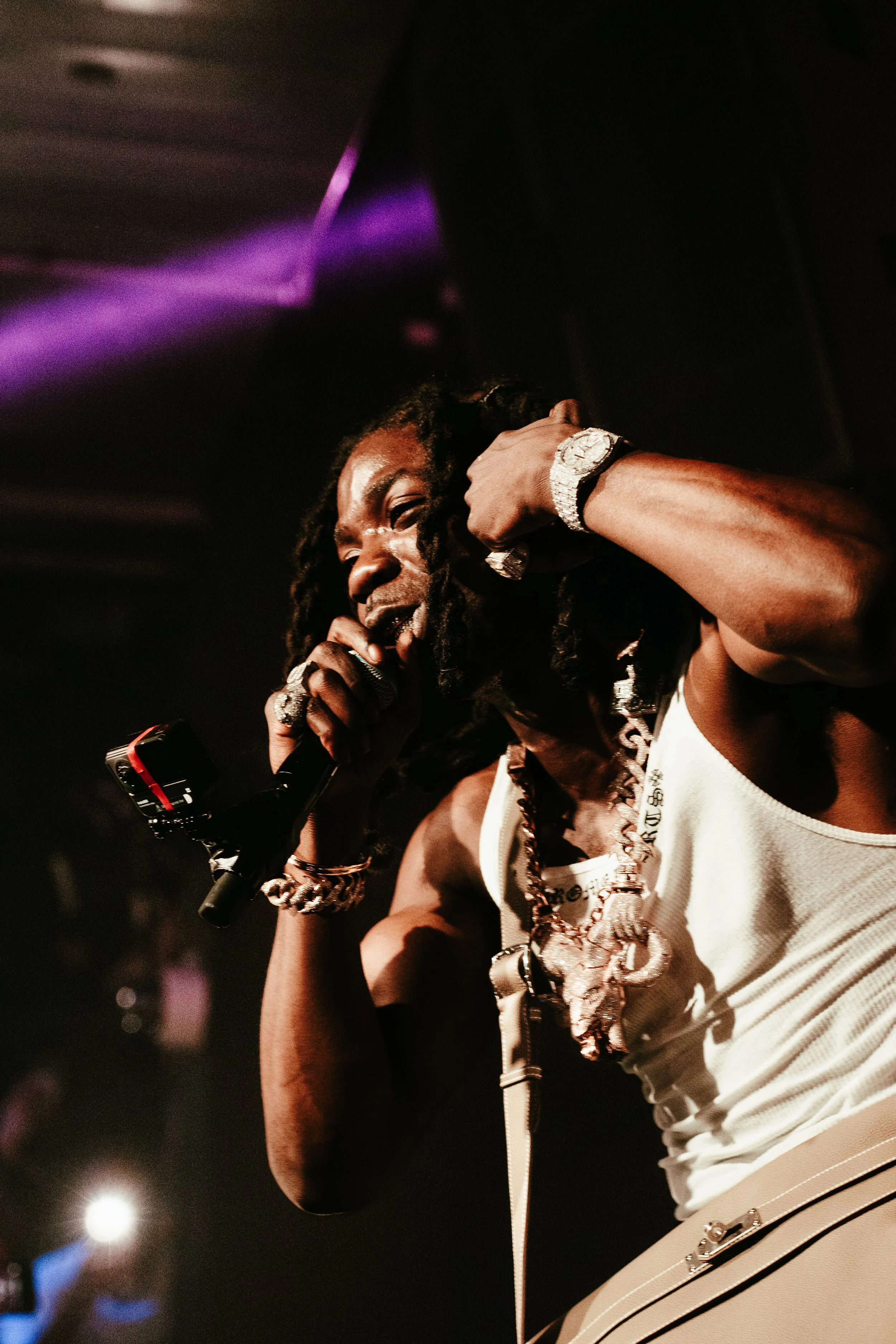 Un artiste en train de chanter dans un micro lors d'une performance, portant des bijoux en or et une tenue blanche, avec une ambiance de scène sombre et colorée.