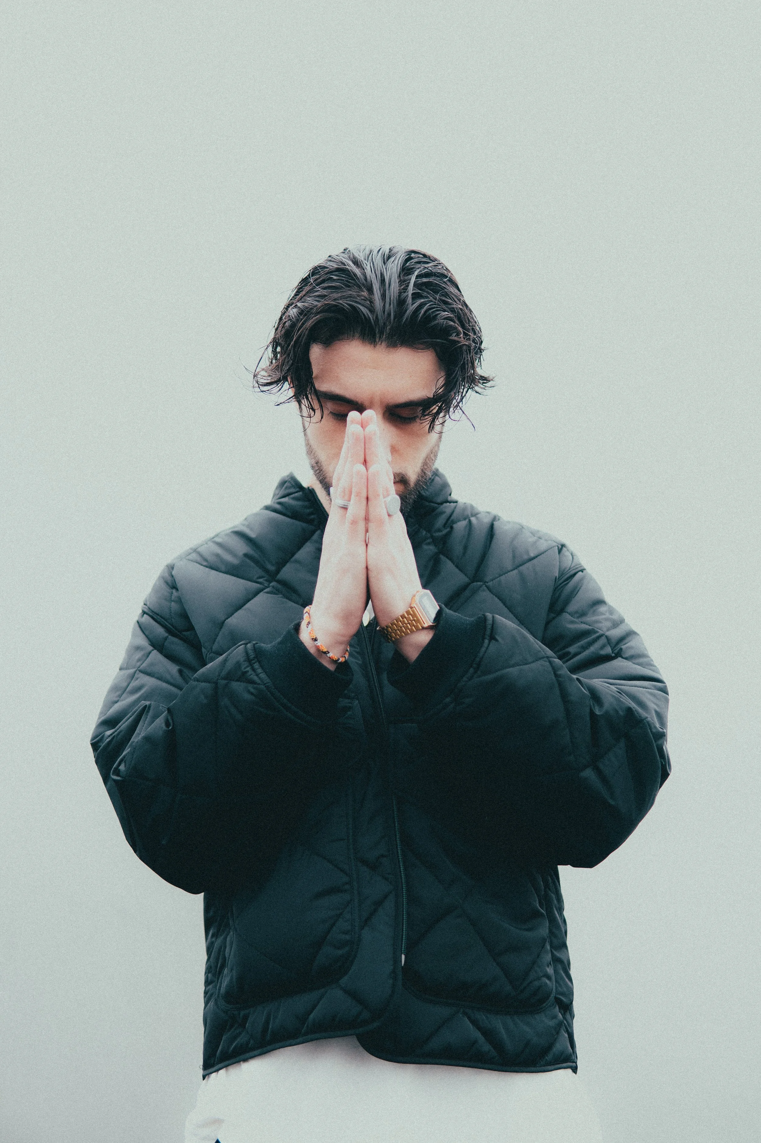 Un jeune homme avec des cheveux noirs, portant un manteau noir, les mains jointes en prière, la tête baissée. Fond gris clair.