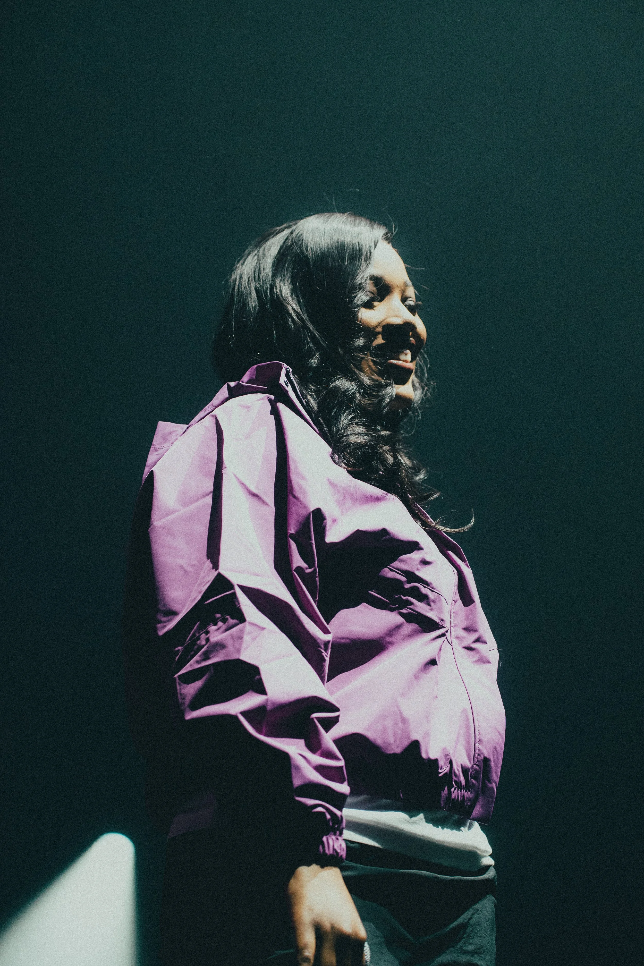 Femme noire avec des cheveux bouclés portant une veste violette, souriant dans un environnement sombre.