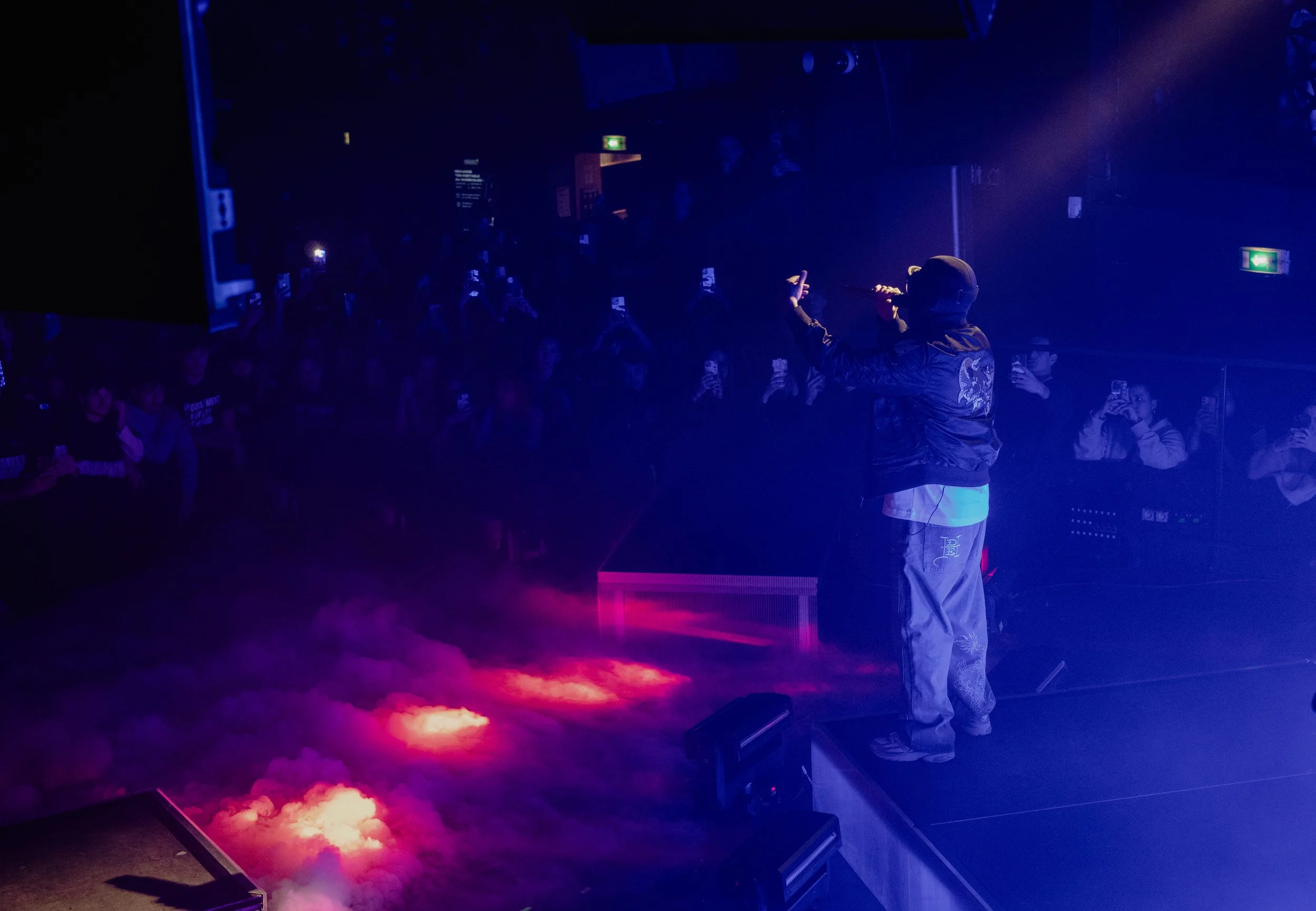 Un artiste sur scène, tenant un microphone, avec une foule de spectateurs en arrière-plan, dans une ambiance sombre et lumineuse avec des effets de lumière colorés et de la fumée au sol.