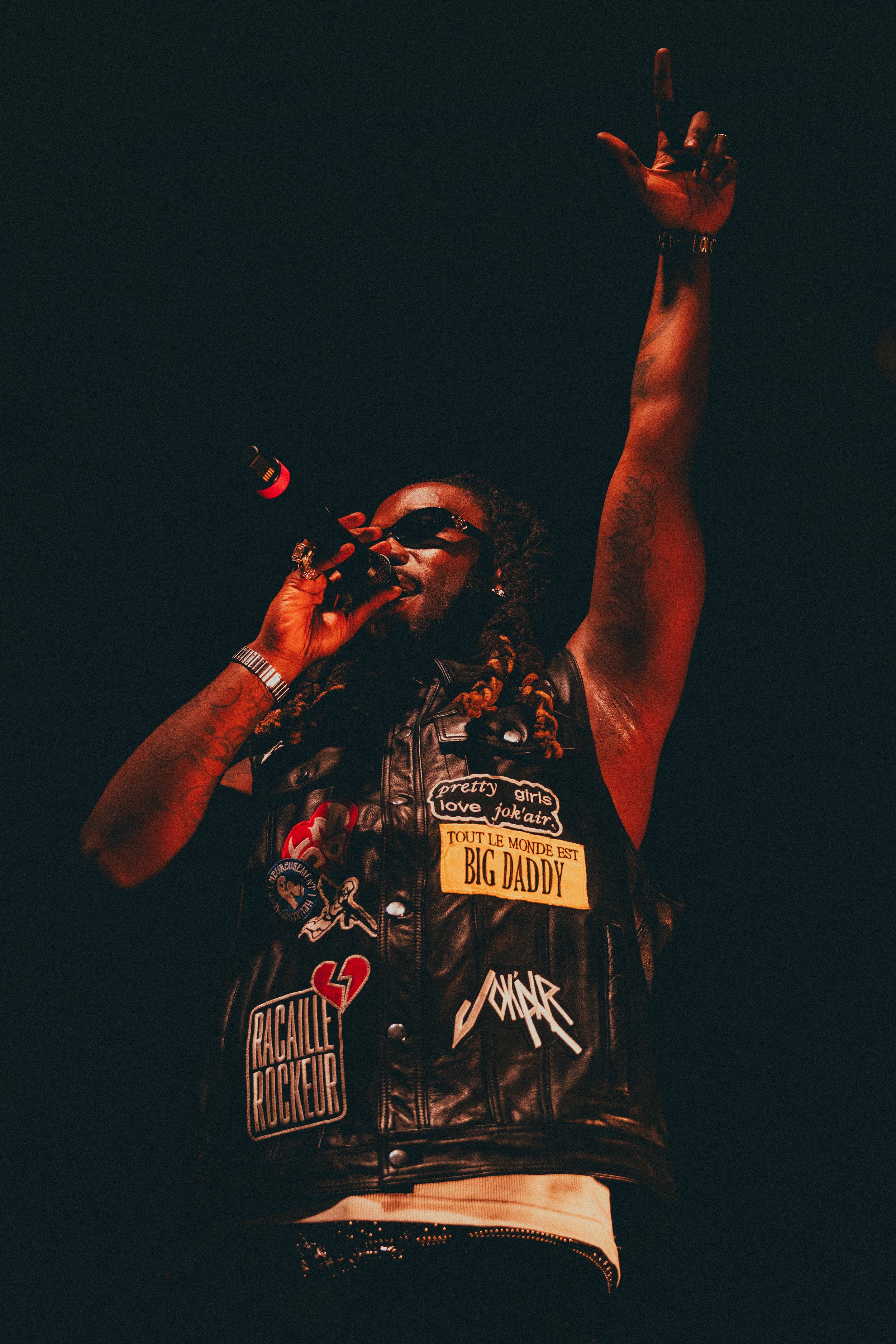 Un homme portant des lunettes de soleil, un gilet en cuir parsemé de patchs, chantant dans un microphone, avec une main levée en l'air, sur scène.