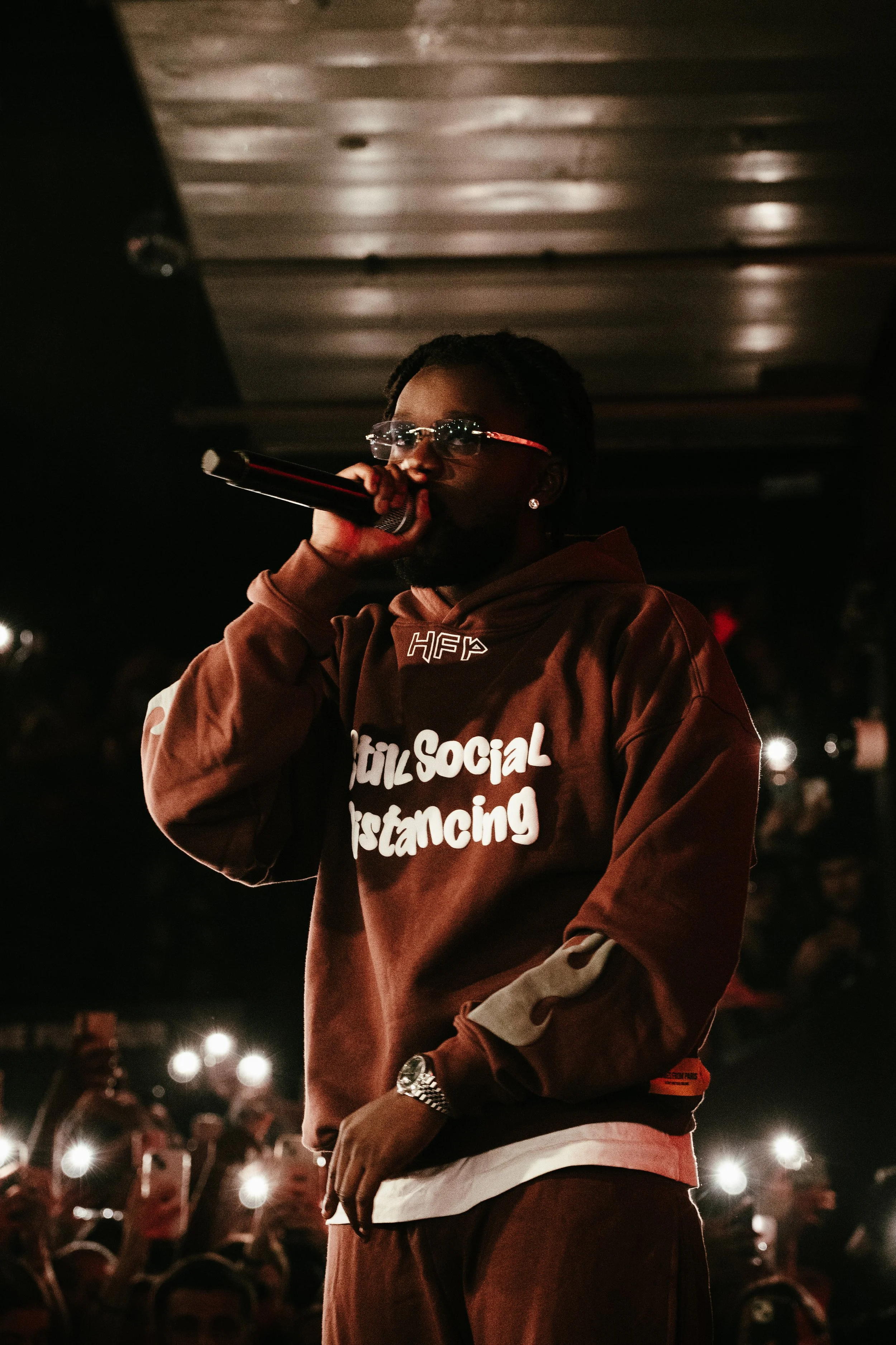 Un artiste portant un sweat marron avec le texte 'Social Distancing' chante dans un micro lors d'un concert. La scène est sombre avec des lumières d'ambiance et des spectateurs en arrière-plan.