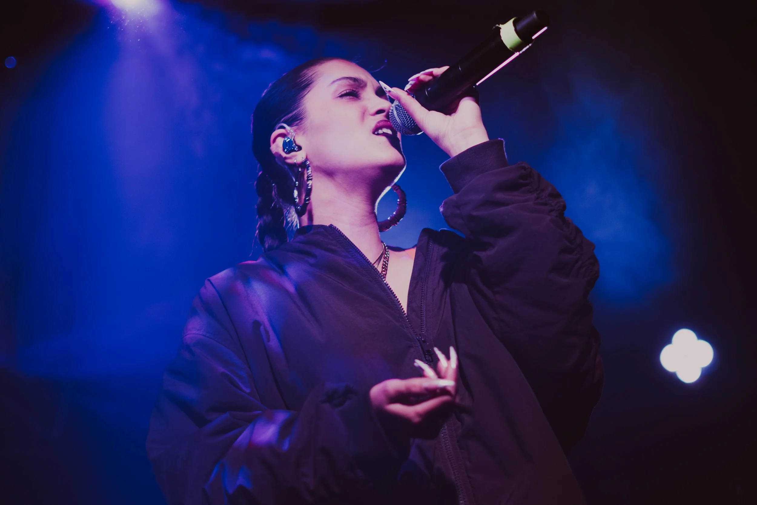 Une femme chante dans un micro, éclairée par des lumières violettes et bleues, portant un blouson noir et des boucles d'oreilles grandes.