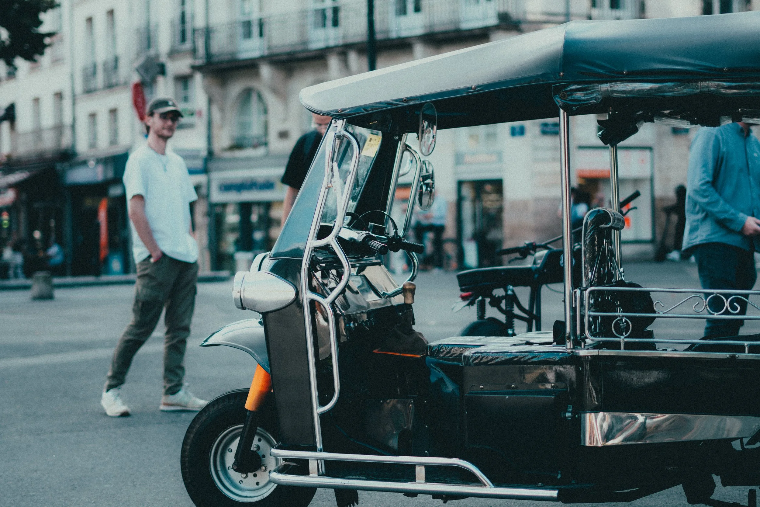 Une voiturette électrique noire et argentée stationnée sur une rue de ville. Deux hommes en arrière-plan, l'un en t-shirt blanc et pantalon beige, et l'autre en veston gris et pantalon noir, marchent ou se tiennent près de la voiturette.