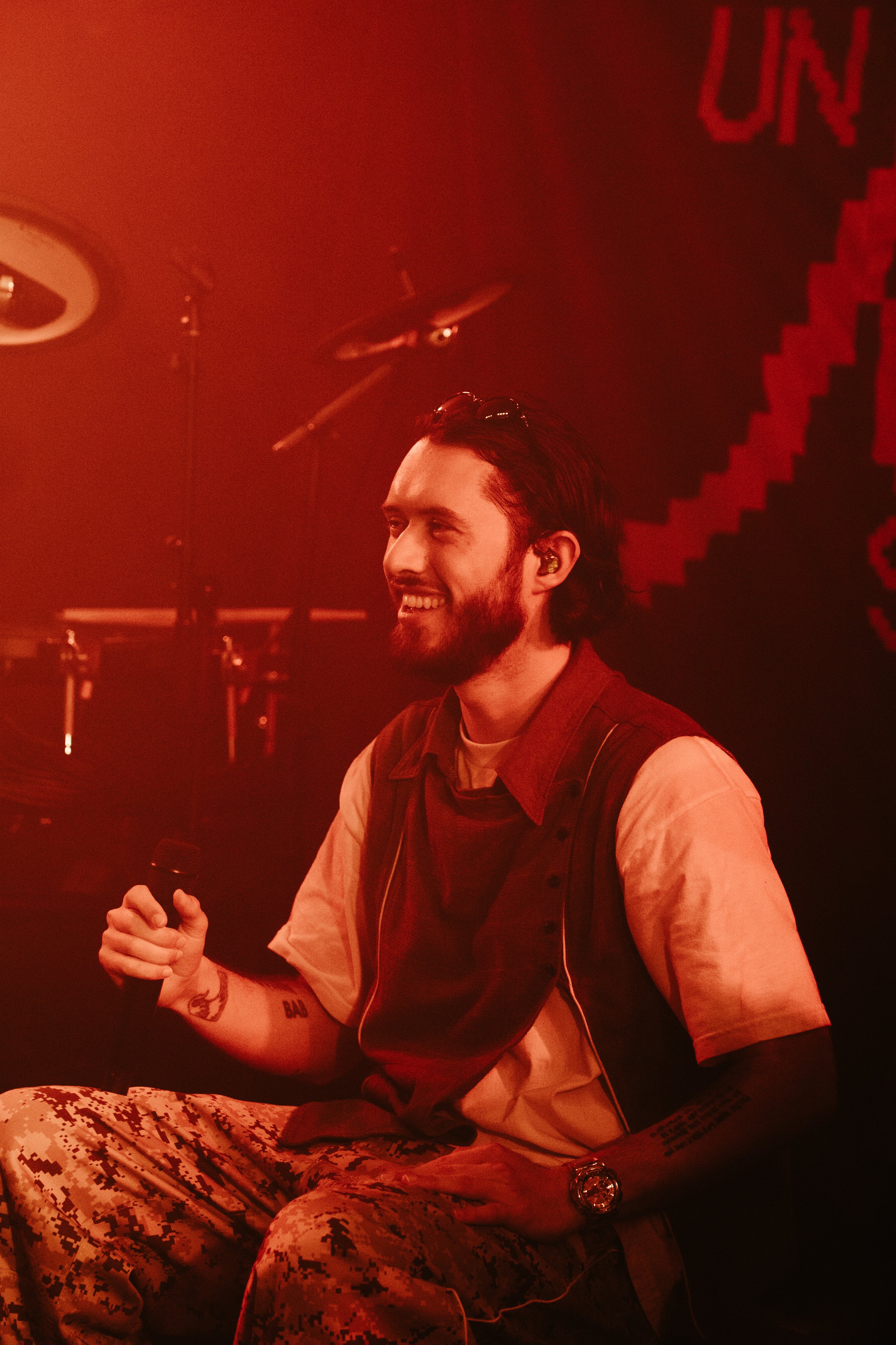 Un homme souriant avec une barbe et des cheveux longs, tenant un microphone, assis dans une ambiance rouge, avec une batterie visible en arrière-plan.