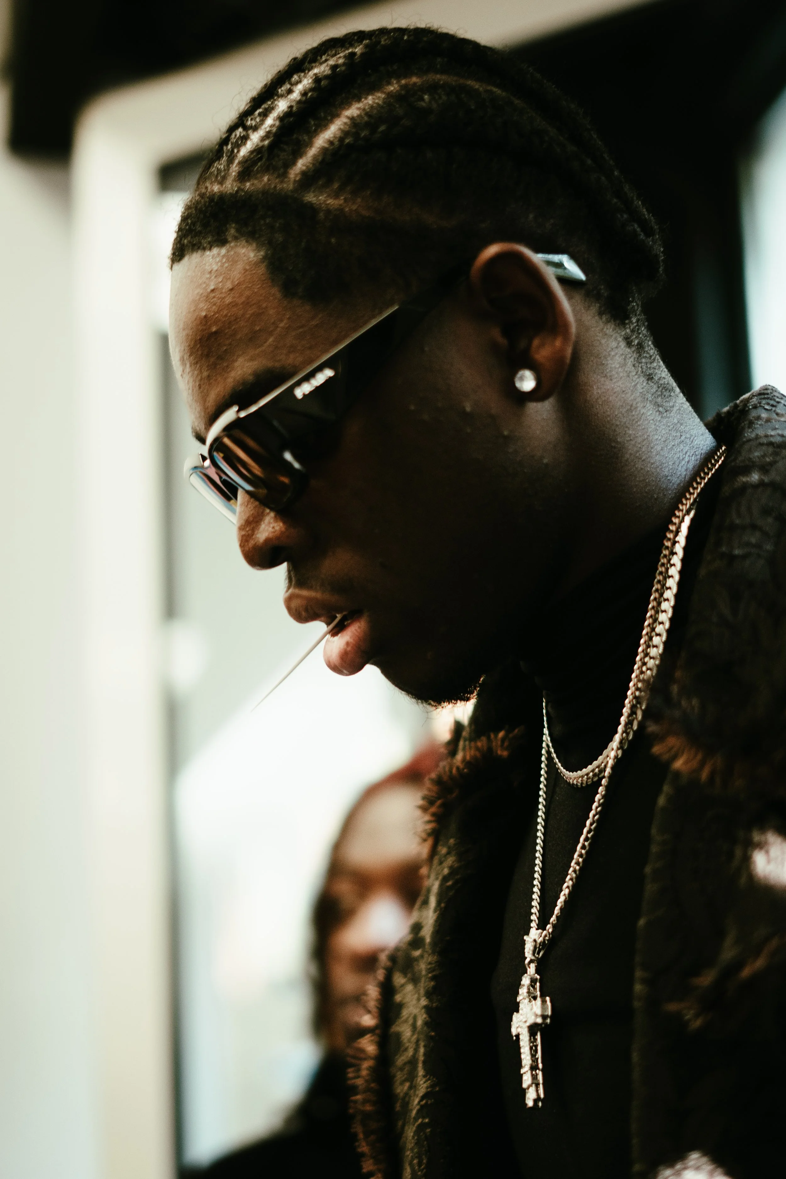 Jeune homme portant des lunettes de soleil noires, des boucles d'oreilles à pierre, un collier avec une croix, habillé d'une veste en fourrure, sortant ou entrant dans un train ou un métro.