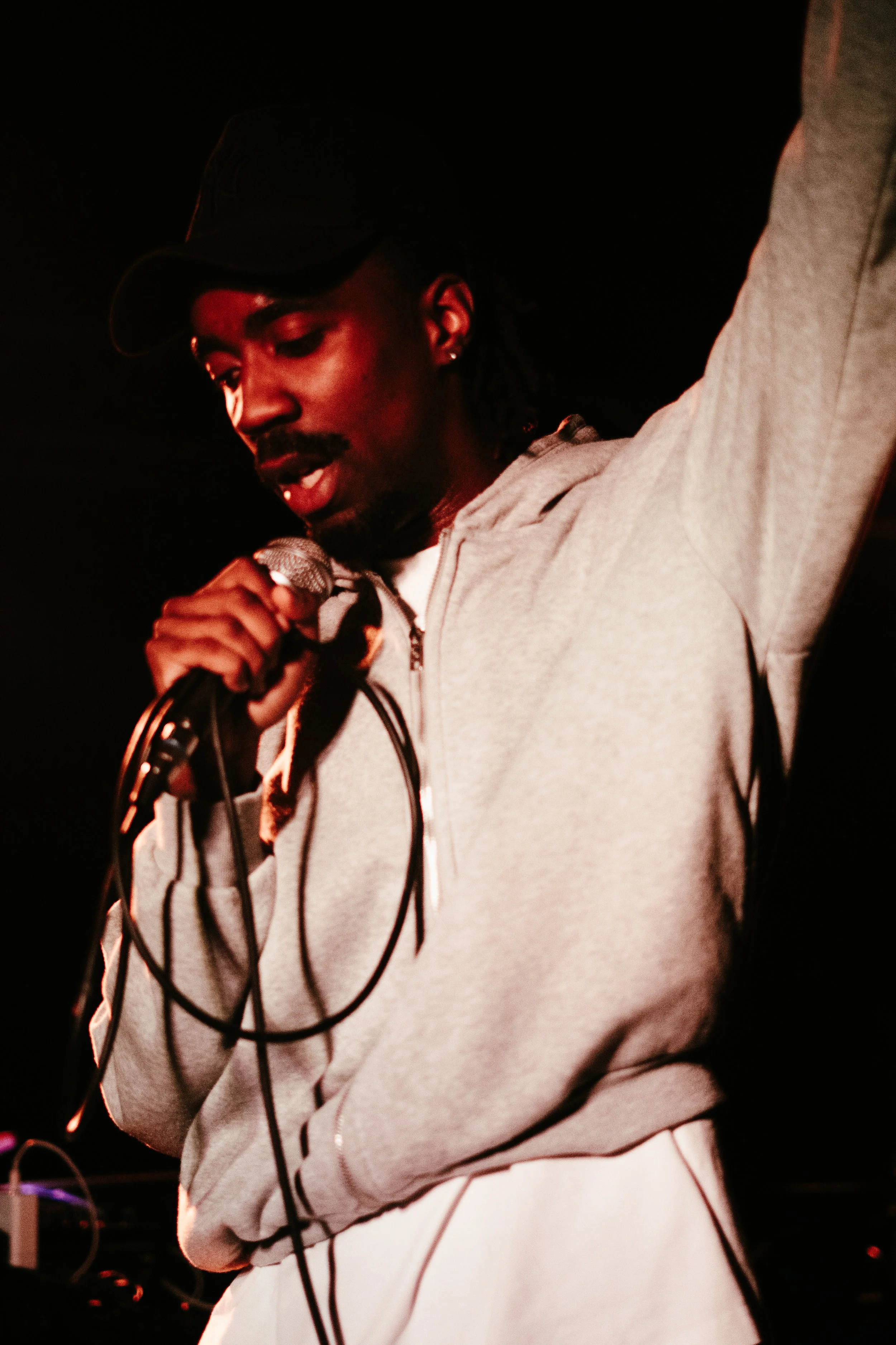 Un jeune homme noir parle ou chante dans un microphone lors d'une performance, portant un sweat à capuche gris et une casquette noire dans un environnement sombre.