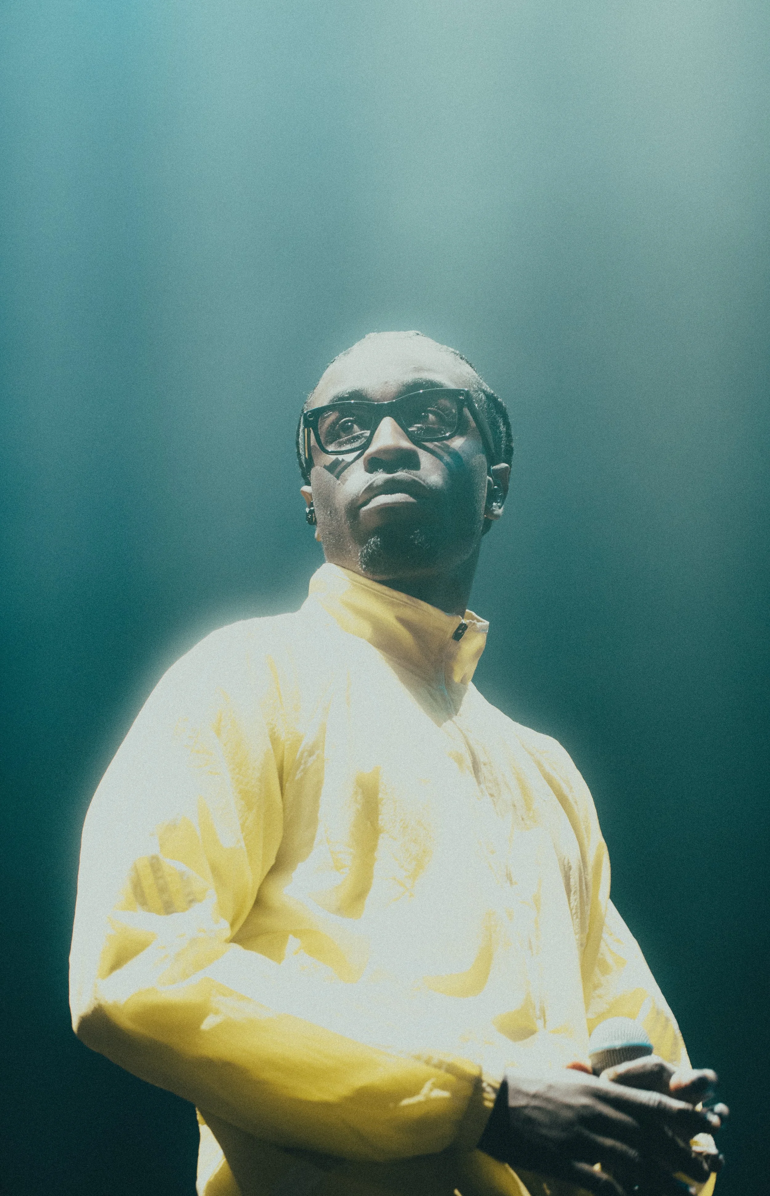 Un homme portant une veste jaune et des lunettes, tenant un microphone, se tient au micro dans une scène sombre.