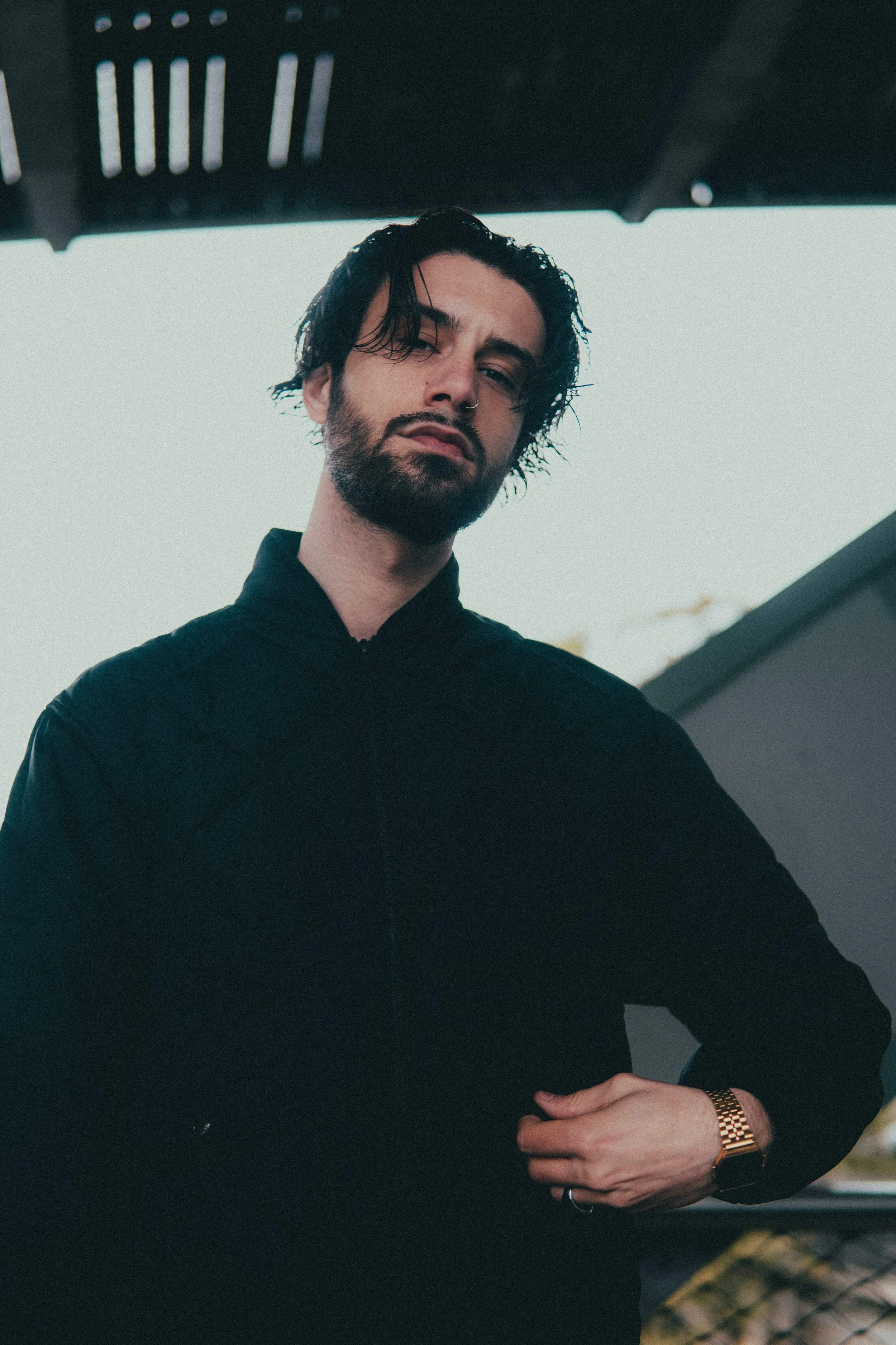 Jeune homme avec barbe, cheveux noirs ondulés, portant un sweat noir et une montre dorée, posant sous un abri.