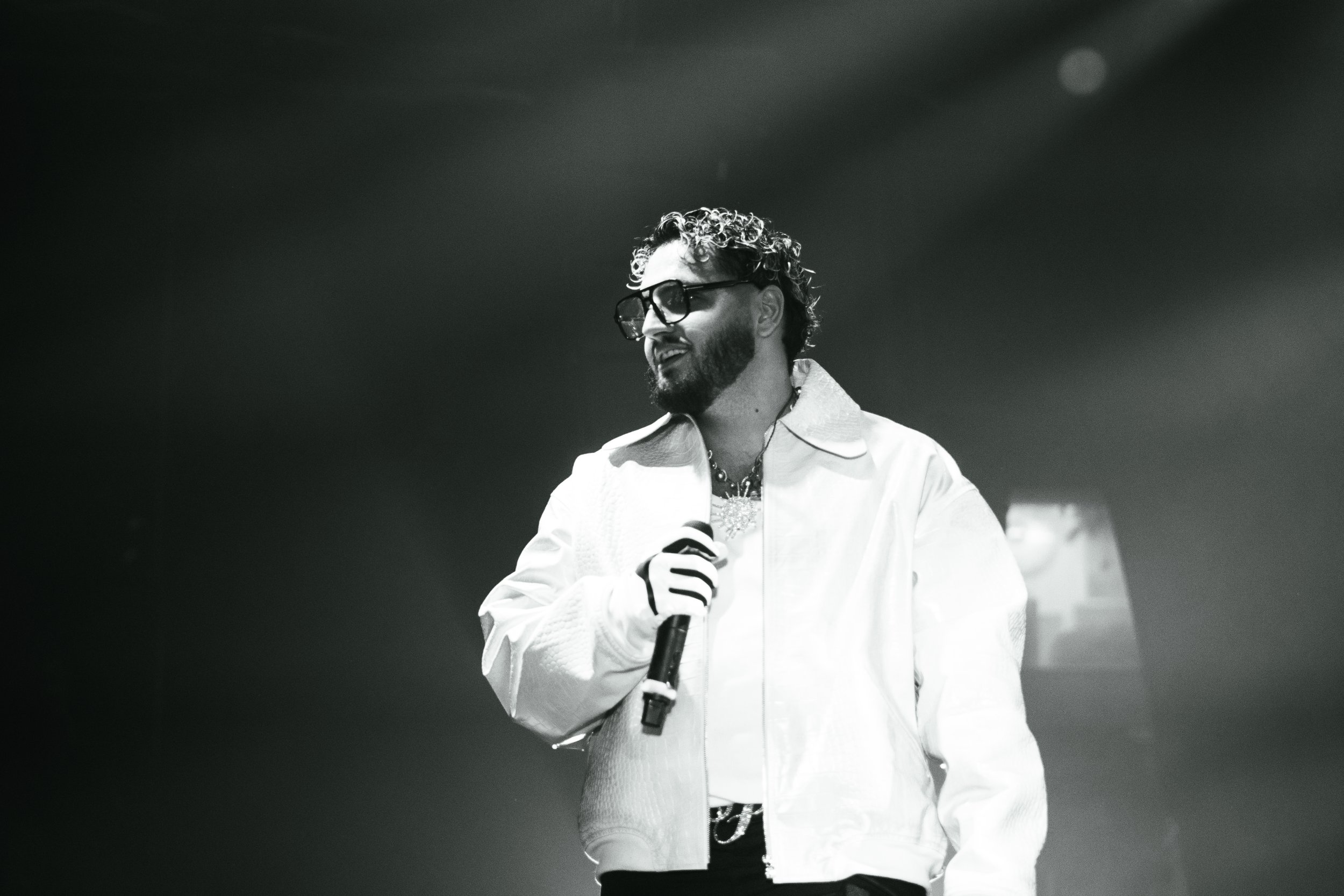 Homme avec lunettes et barbe, tenant un microphone, en train de parler ou chanter, dans une lumière tamisée.