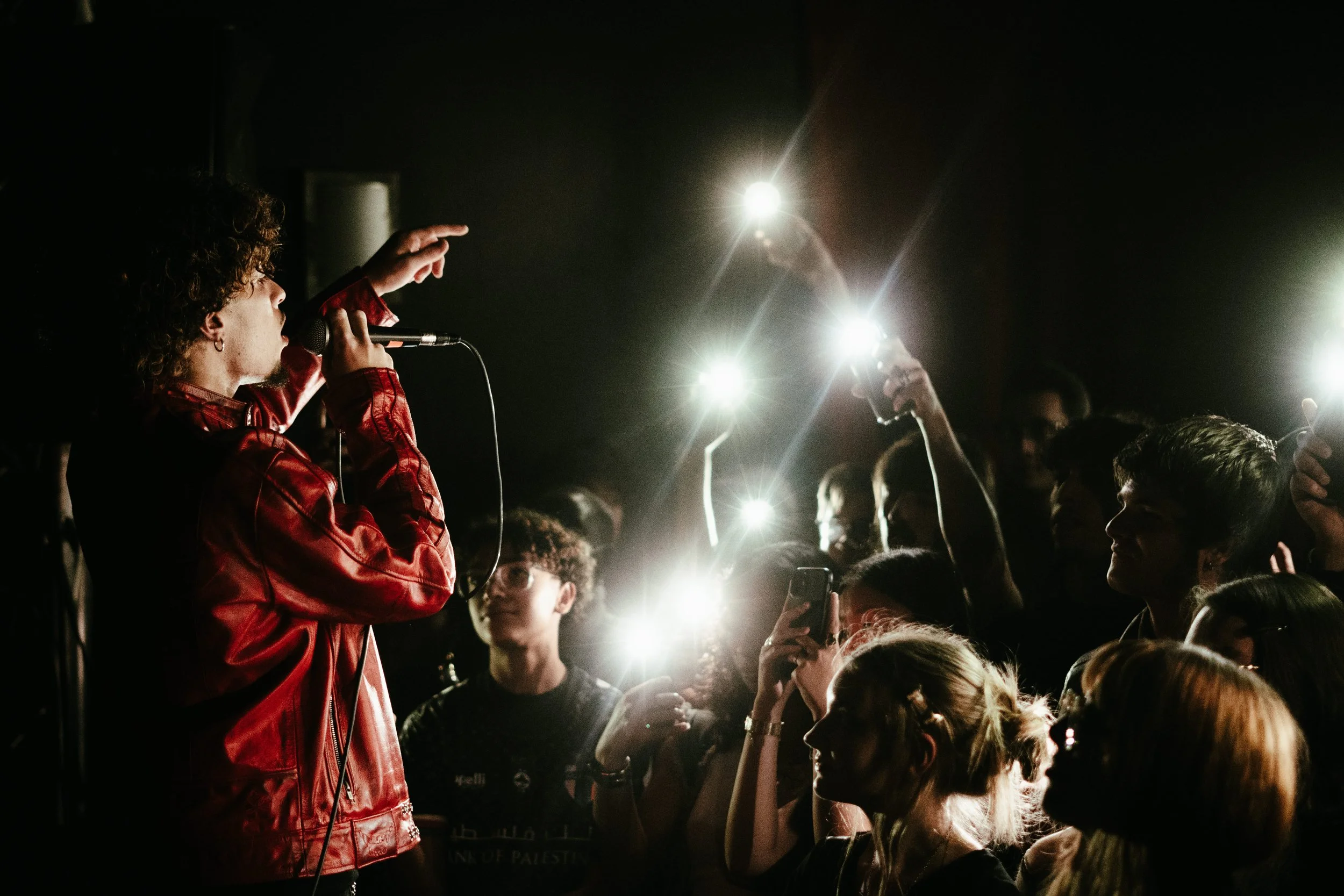 Un chanteur avec une veste en cuir rouge lors d'un concert, entouré d'un public qui l'éclaire avec leurs téléphones portables.