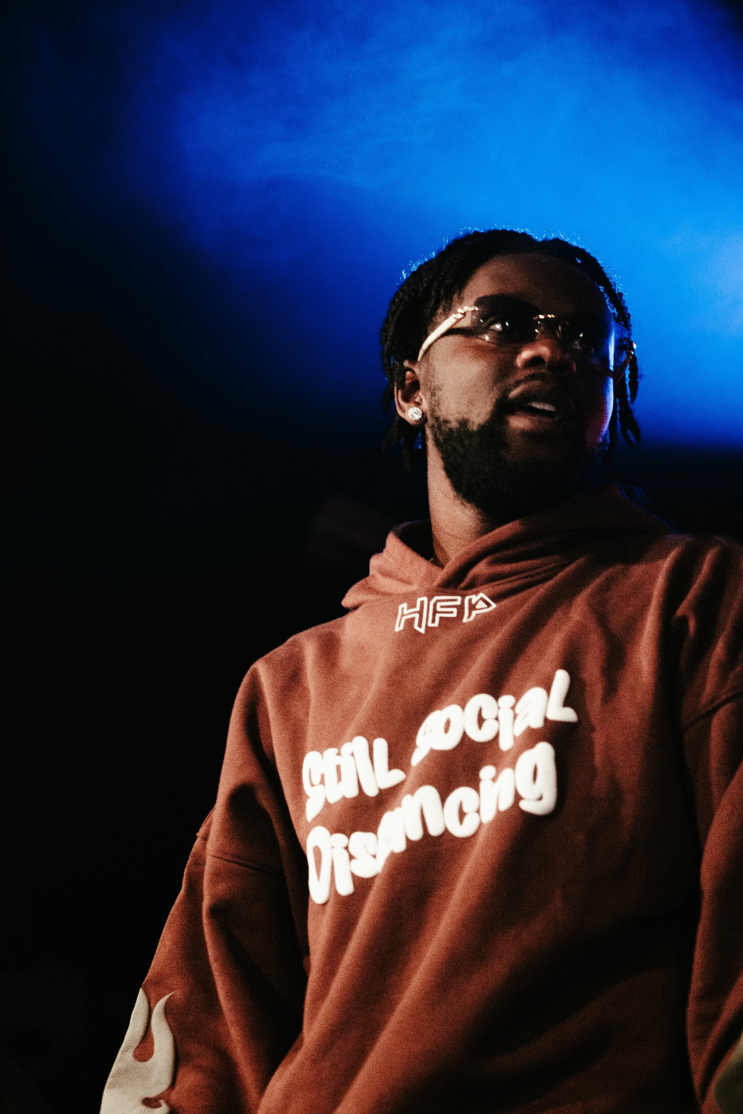 Un homme portant un sweat à capuche marron avec l'inscription « still social distancing » en blanc, portant des lunettes et des boucles d'oreilles, sous un éclairage bleu.