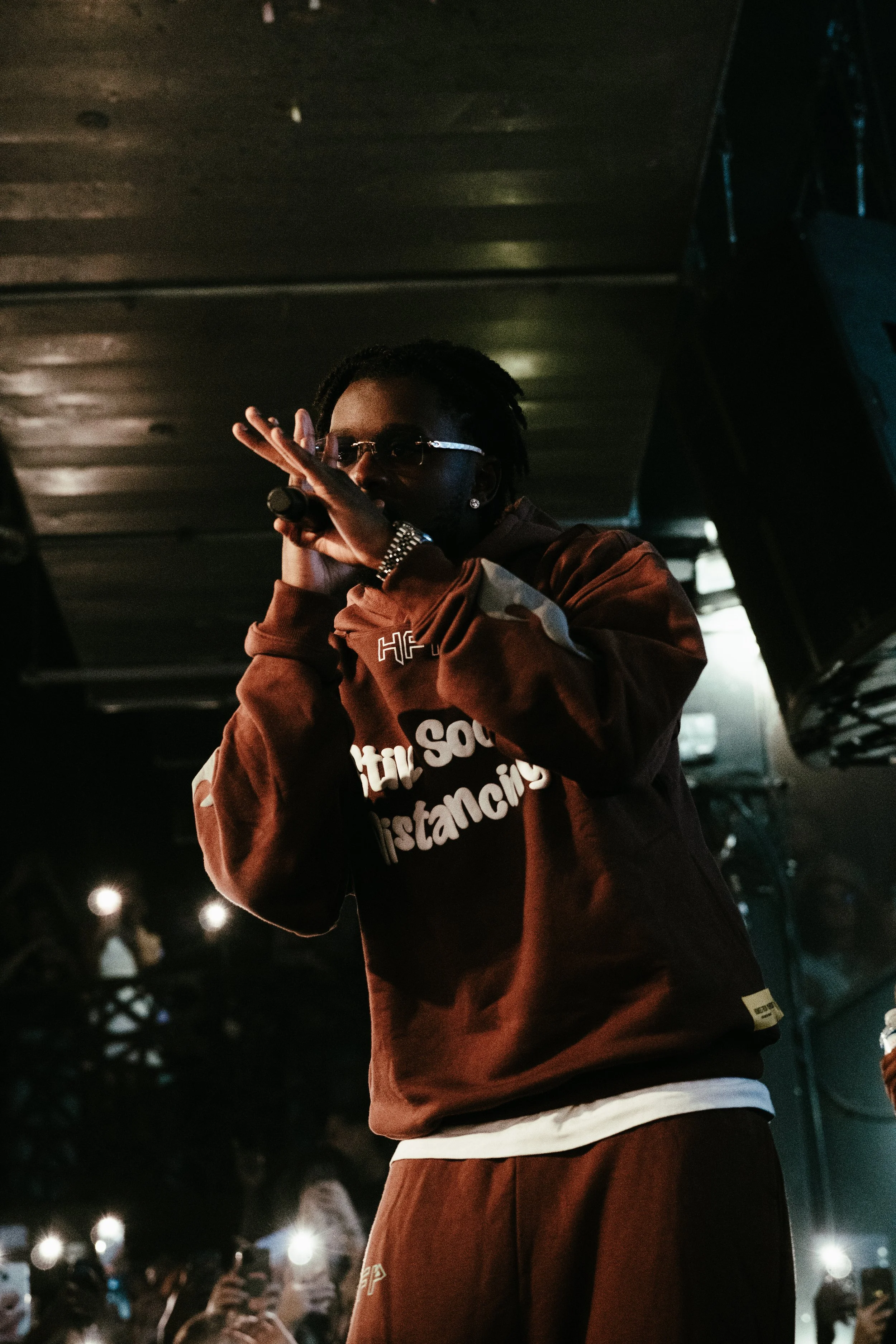 Un artiste de rap ou chanteur performe sur scène, portant une veste marron avec un message en français, avec des écouteurs dans les oreilles, en pleine présentation devant un public.